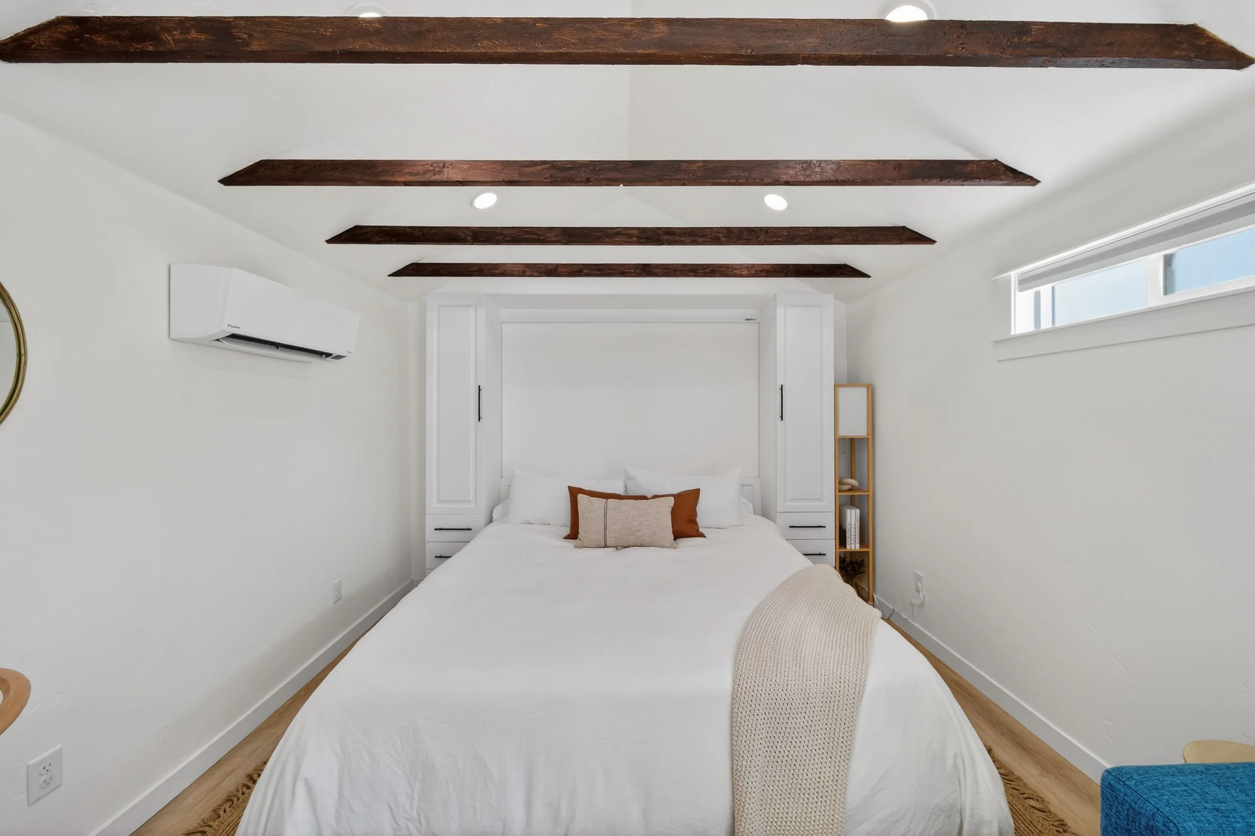 A bedroom with a white bed, wooden ceiling beams, and a window high on the wall, featuring minimal decor.