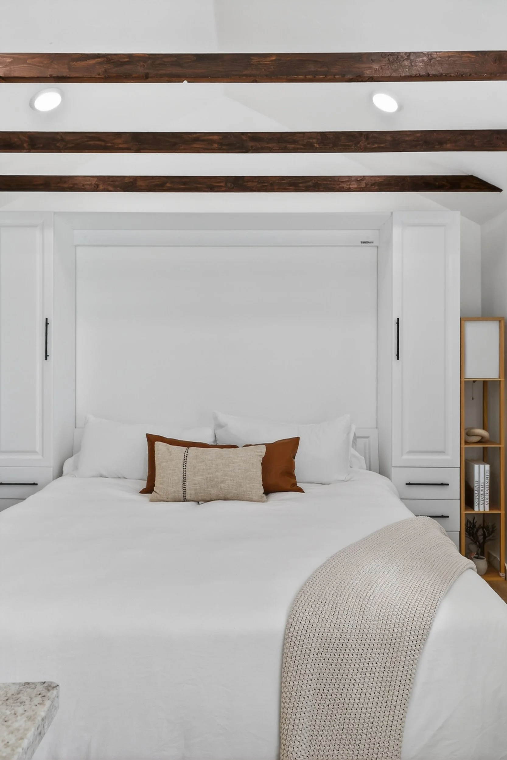 A neatly made bed with white bedding, two white pillows, and two decorative pillows in shades of brown and beige, in a modern bedroom with white walls and dark wooden ceiling beams.