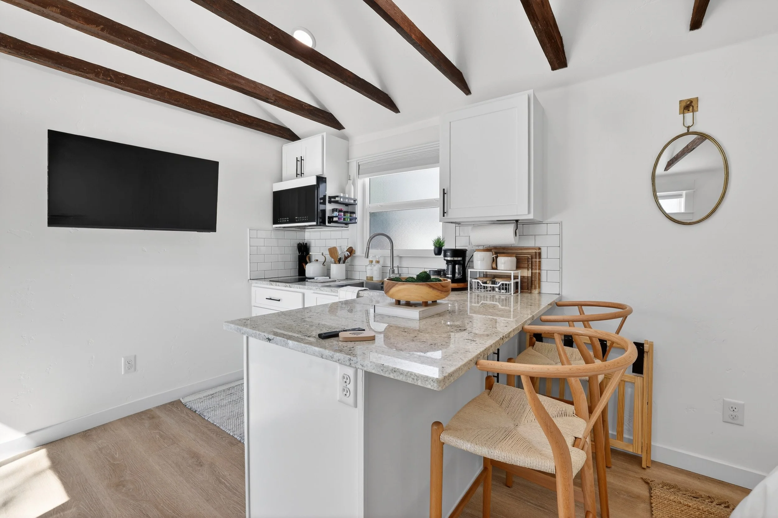 Modern kitchen with white cabinets, granite countertop, subway tile backsplash, wooden ceiling beams, and two wooden chairs at the breakfast bar. There is a small round mirror on the wall.