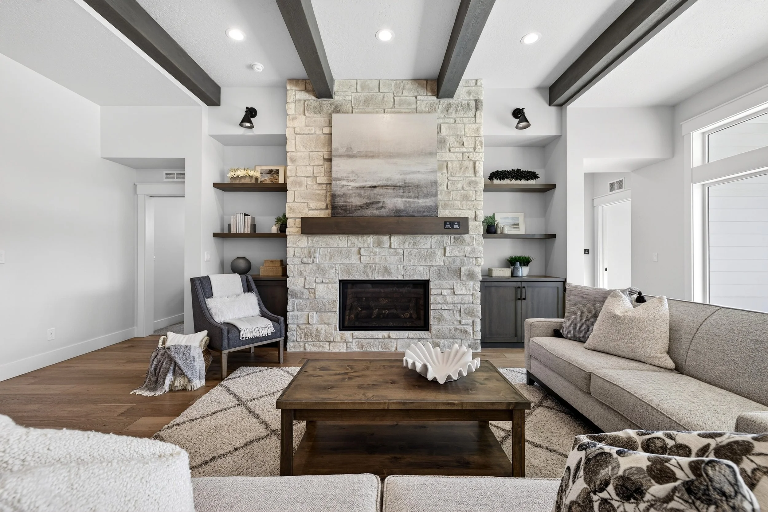 Living room with a stone fireplace, beige sofa, wooden coffee table, armchair with throw blanket, built-in shelves, large windows, and modern decor.