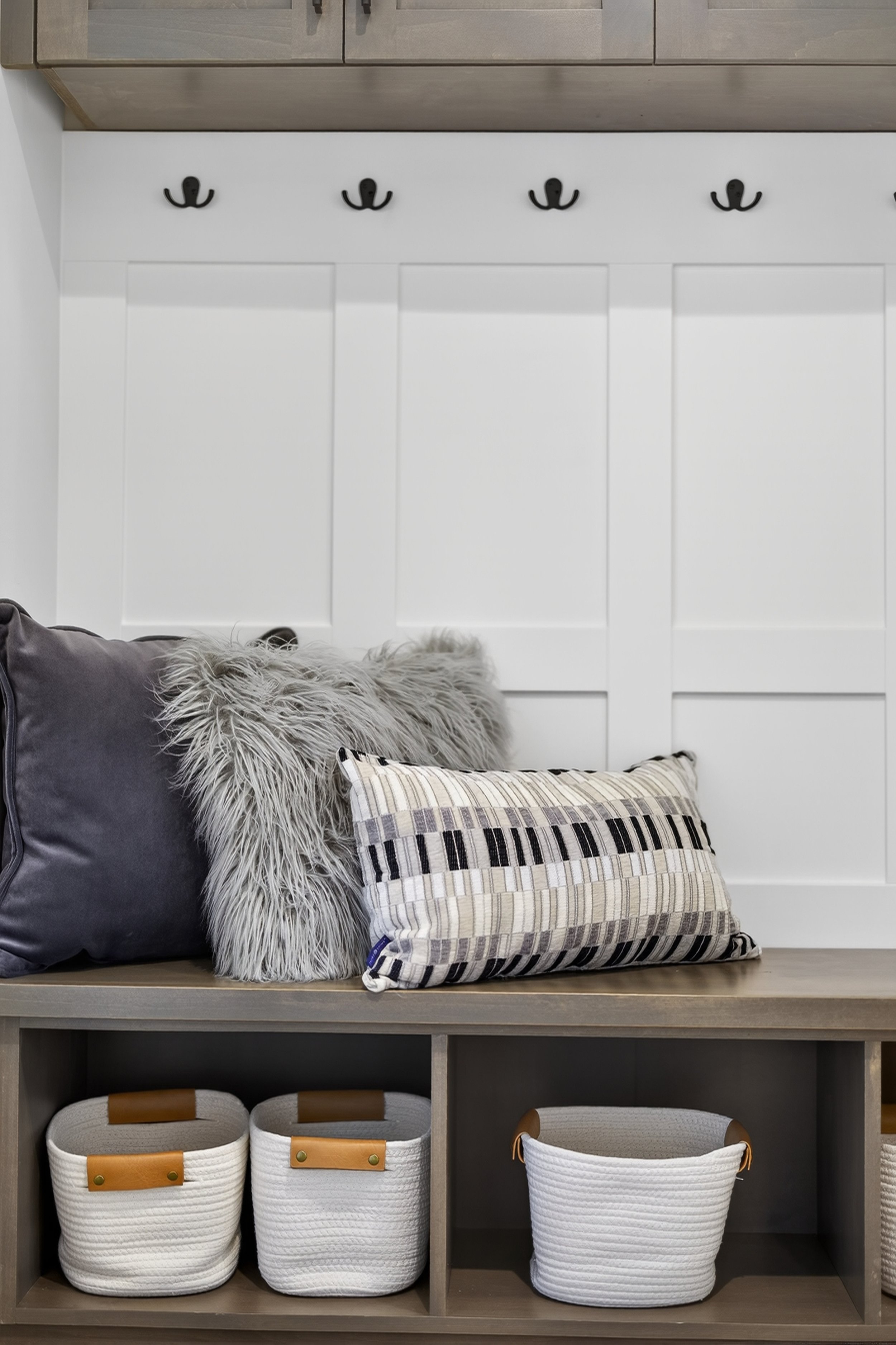 A wooden bench with three decorative pillows, and storage baskets underneath. Above, four black hooks on a white paneled wall.
