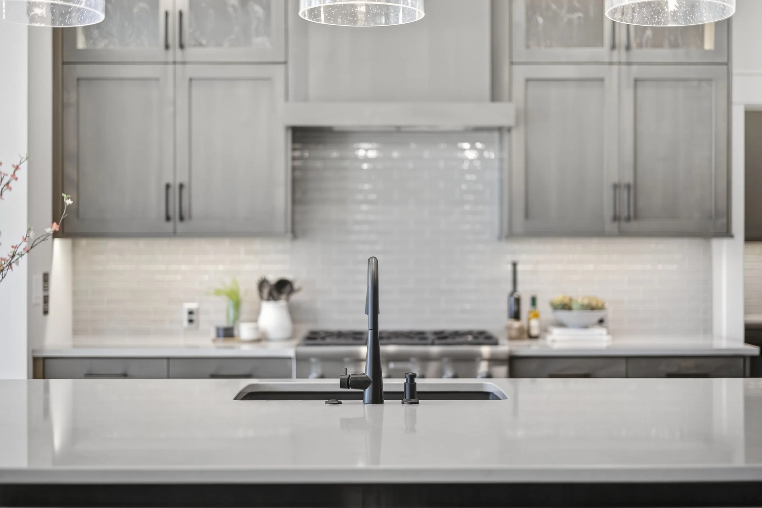 Modern kitchen with a white countertop, black sink faucet, and gray cabinets. Items on the countertop include a white pitcher, bottles, and a bowl of fruit.