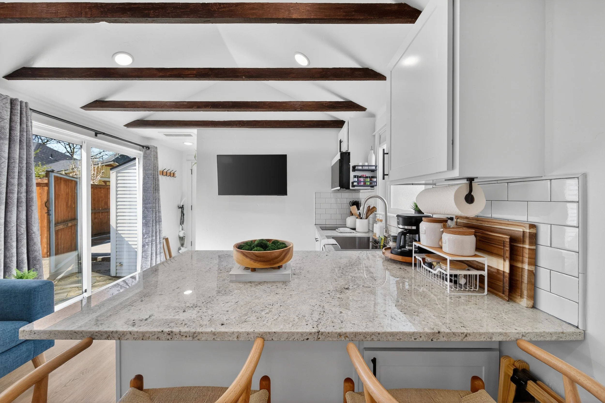 Kitchen with a large beige granite island, wooden beams on the ceiling, white cabinets, and a sliding glass door leading outside.