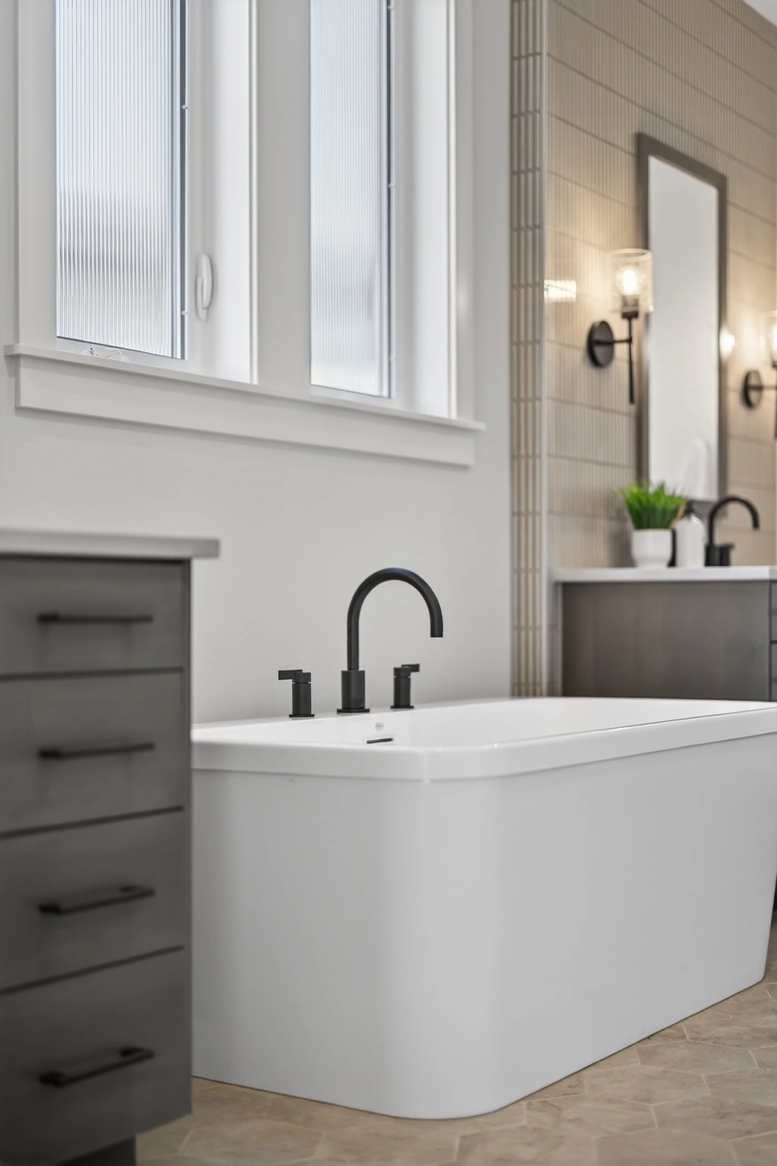 Modern bathroom with a white bathtub, black fixtures, gray cabinets, and plants.