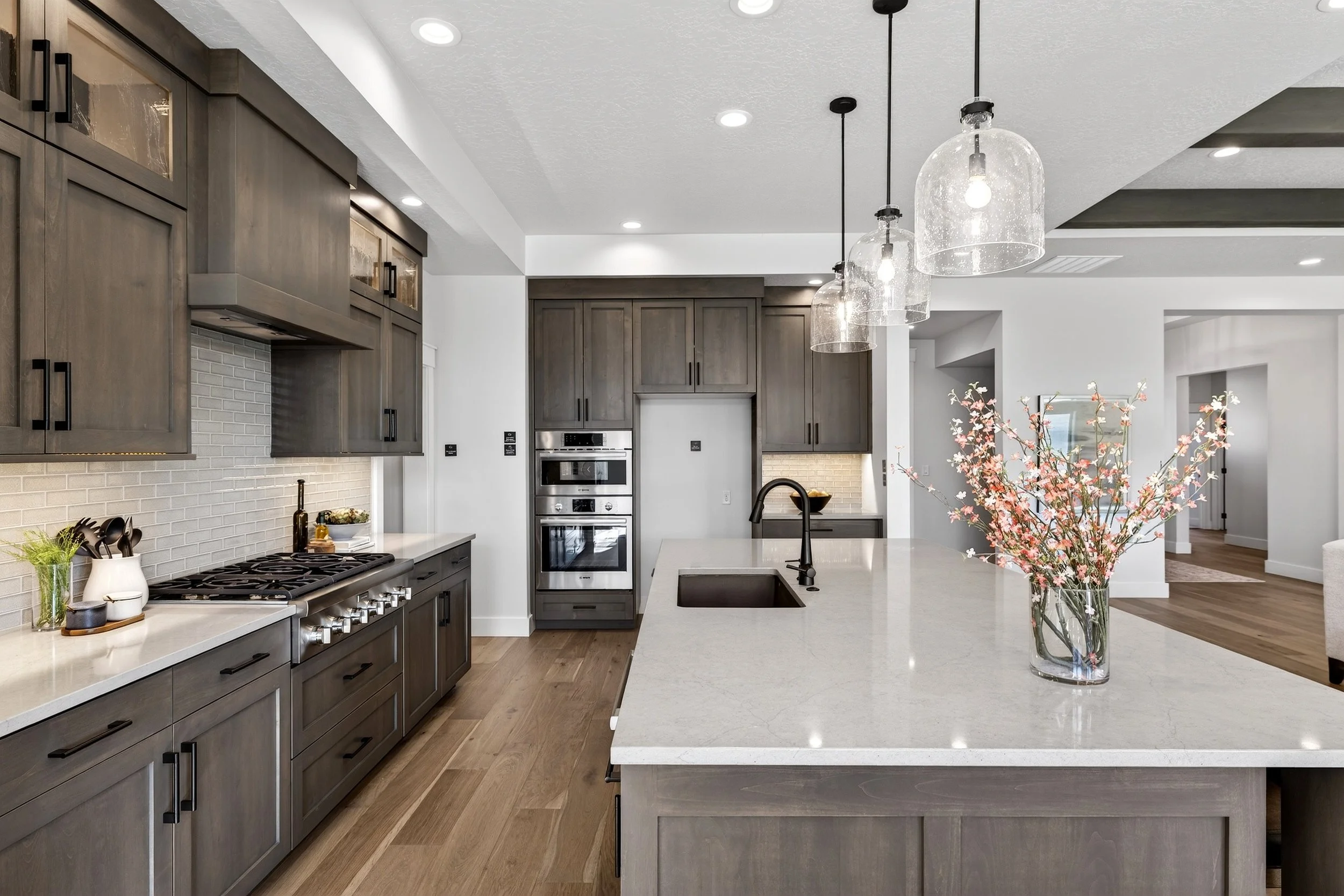 Modern kitchen with gray cabinets, white countertops, and a large kitchen island with a sink and a black faucet. There is a vase with pink flowers on the island, hanging pendant lights above, and wooden flooring.