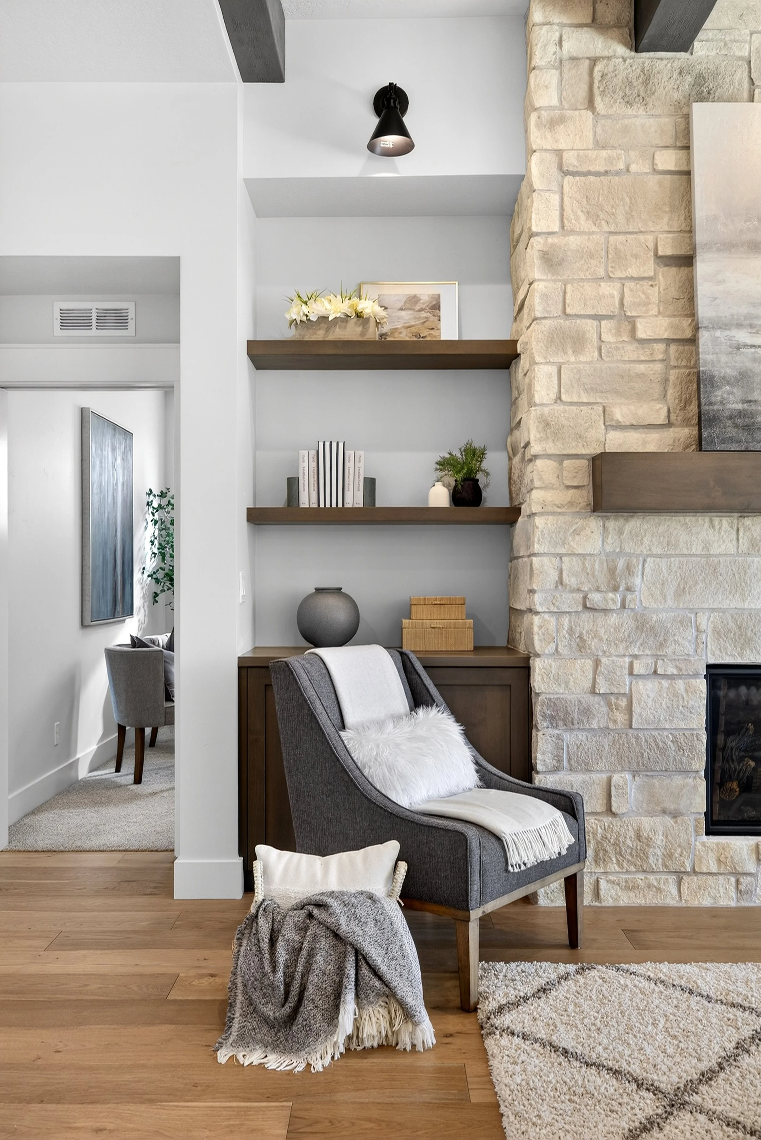 Corner of a living room with a gray armchair, fluffy white pillow, and blanket, next to a built-in wooden cabinet and open shelving with decorative items, a stone fireplace, hardwood floors, and a doorway leading to a dining area.
