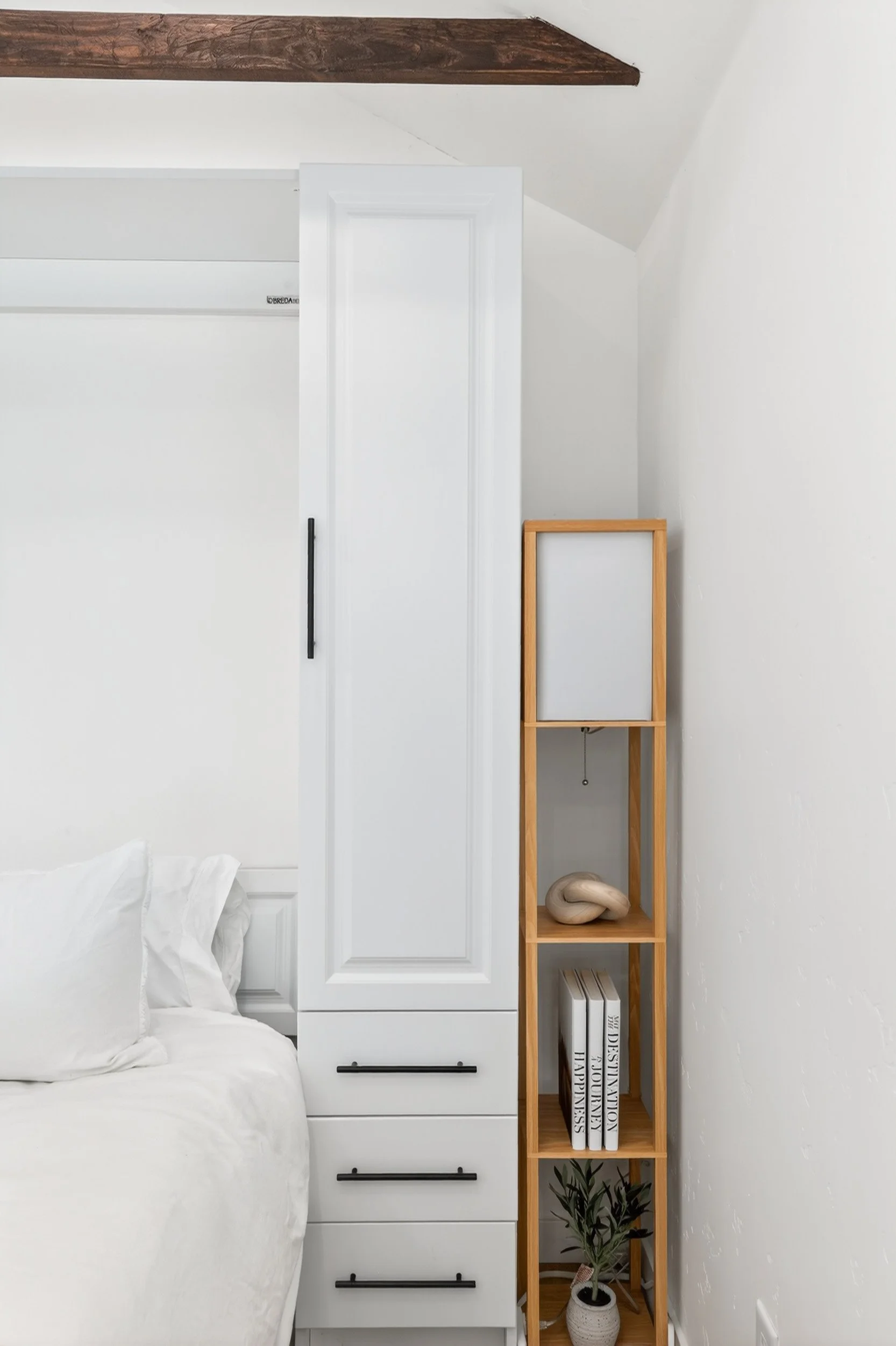 A bedroom corner featuring a white bed with white pillows, a tall white wardrobe with black handles, a wooden shelving unit with decorative items and books, and a green potted plant.