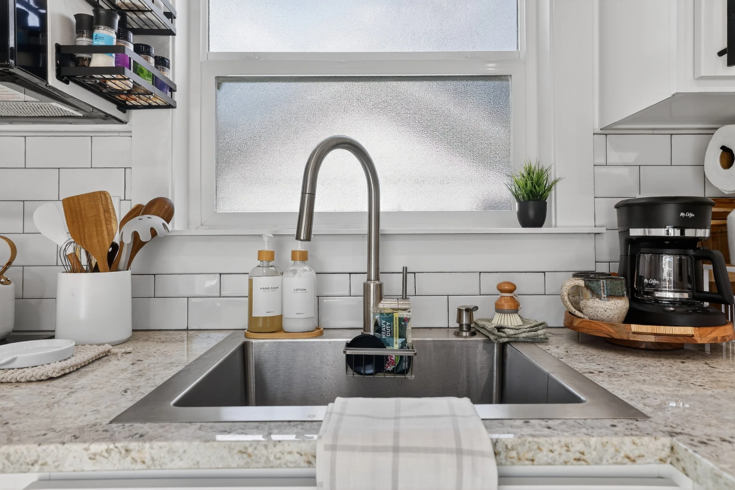 Kitchen sink area with faucet, soap and lotion bottles, dish soap, a coffee maker, paper towels, a small potted plant, and utensils in a white container, with a window in the background.