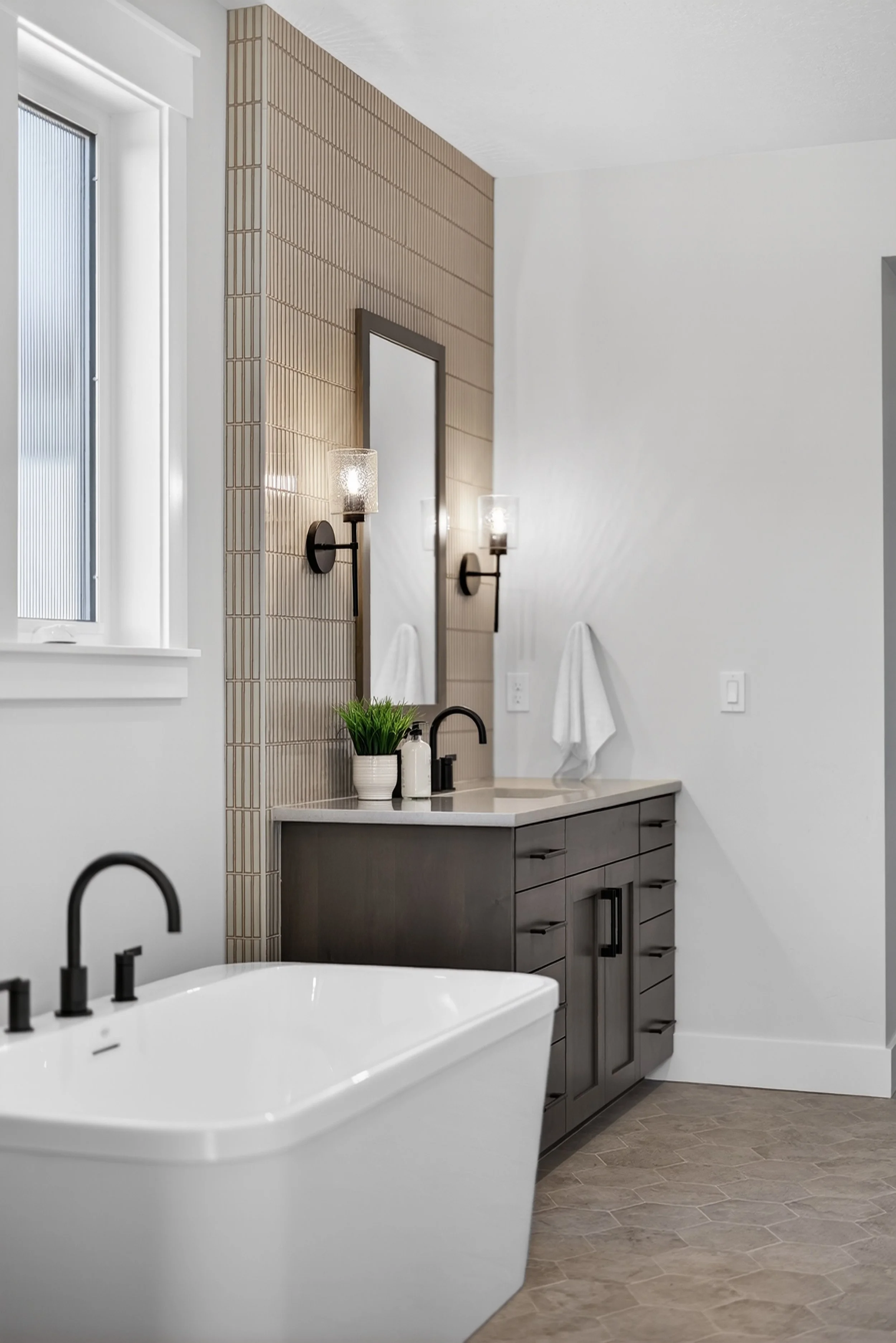 Modern bathroom with a bathtub, a vanity with a mirror, wall-mounted lights, a window, a towel, and a small potted plant.