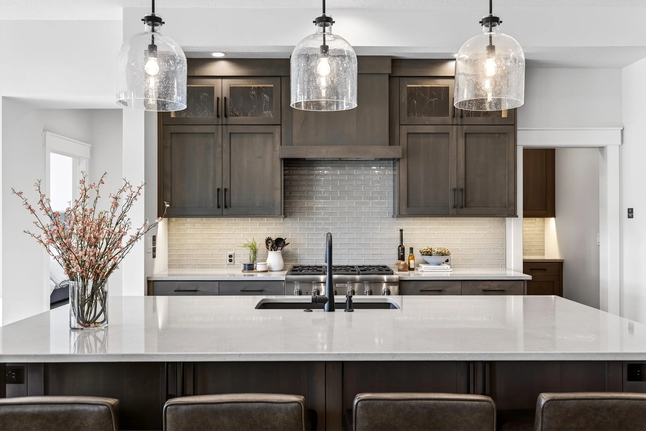 Modern kitchen with dark wood cabinets, white tiled backsplash, white countertop, and three hanging glass pendant lights. The island has a sink, a vase with pink flowers, and bar stools.