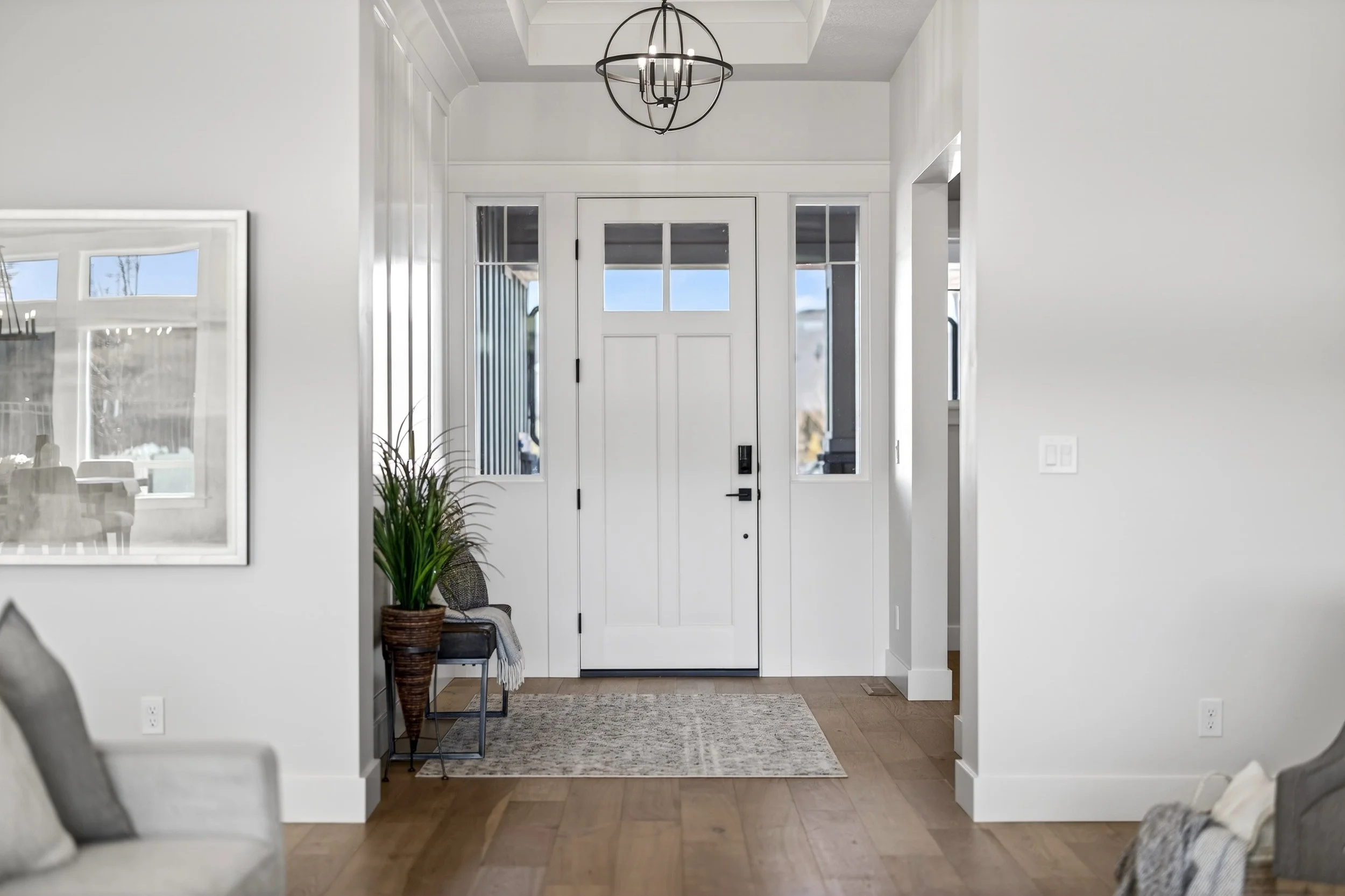 Front entryway of a modern home with a white door, surrounded by windows, a potted plant, a bench with a throw blanket, and a chandelier overhead.