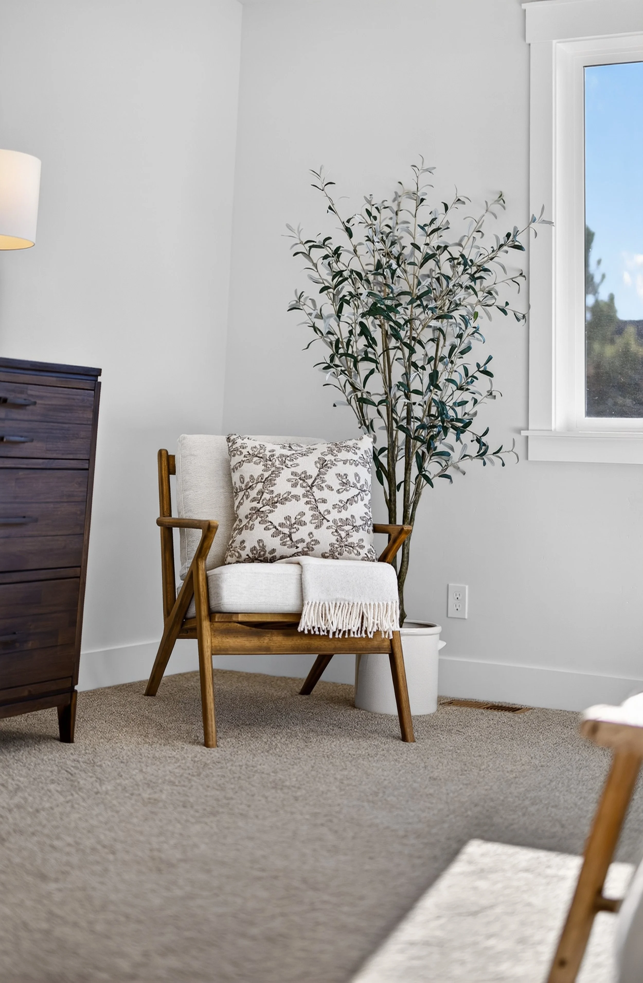 A cozy room corner with a white armchair adorned with a patterned pillow and a fringed blanket, a large potted plant with dark green leaves, a wooden dresser, and a window showing a blue sky with a few clouds.