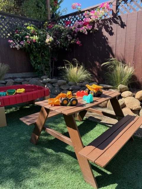 Backyard with social play area, including a wooden picnic table with toys on it, a paddle boat, and a garden with flowers and ornamental grasses, fenced with lattice and brown panels.