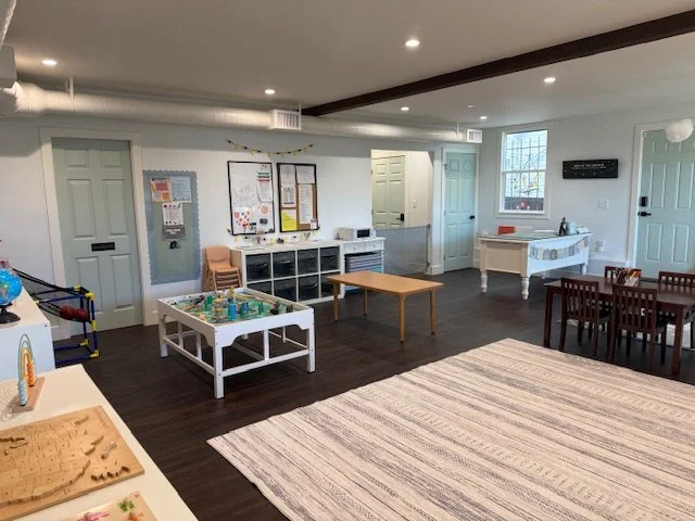 Indoor playroom or classroom with small chairs, tables, a white piano, a rug, and various toys and items for children.