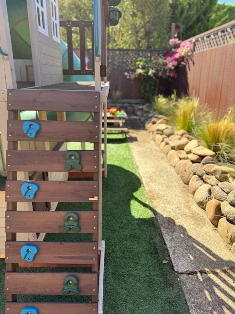 Backyard with a wooden play structure, climbing holds, artificial grass, and a stone walking path with garden plants and flowers.