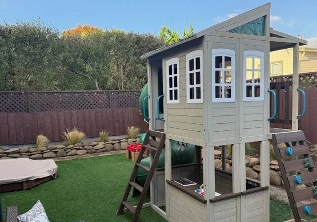 Backyard with a wooden playhouse featuring a slide, small stairs, and a sandbox, on artificial grass with a fence and trees in the background.