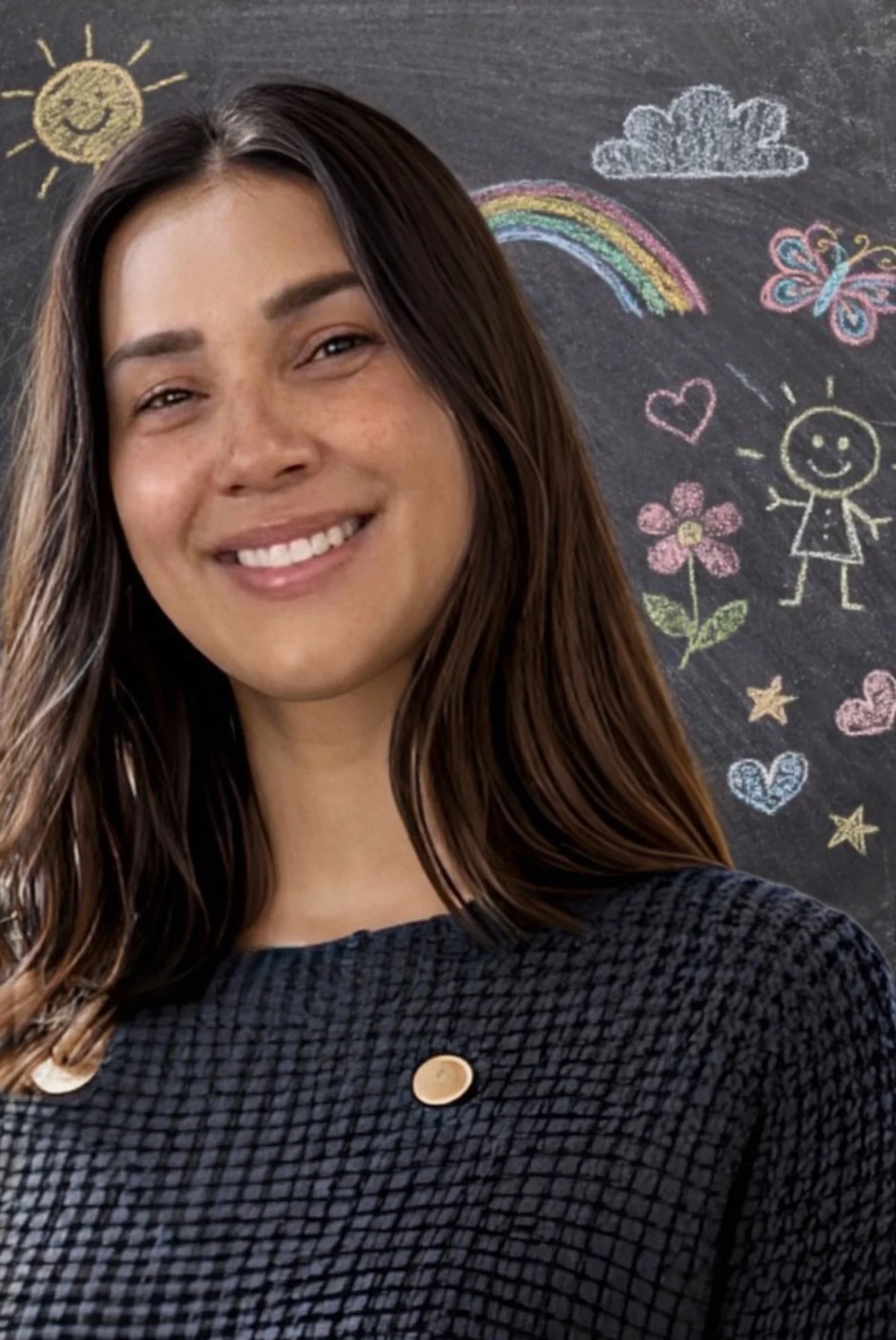 A smiling woman with long brown hair standing in front of a blackboard decorated with colorful chalk drawings of a sun, rainbow, cloud, butterfly, flower, hearts, stars, and a girl.