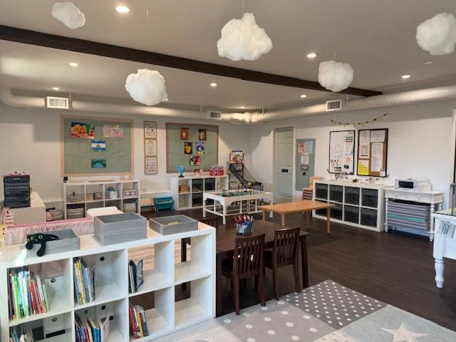 Classroom decorated with cloud-shaped paper lanterns hanging from ceiling, with white walls, shelves, and children's furniture, including a bookshelf, tables, and chairs.