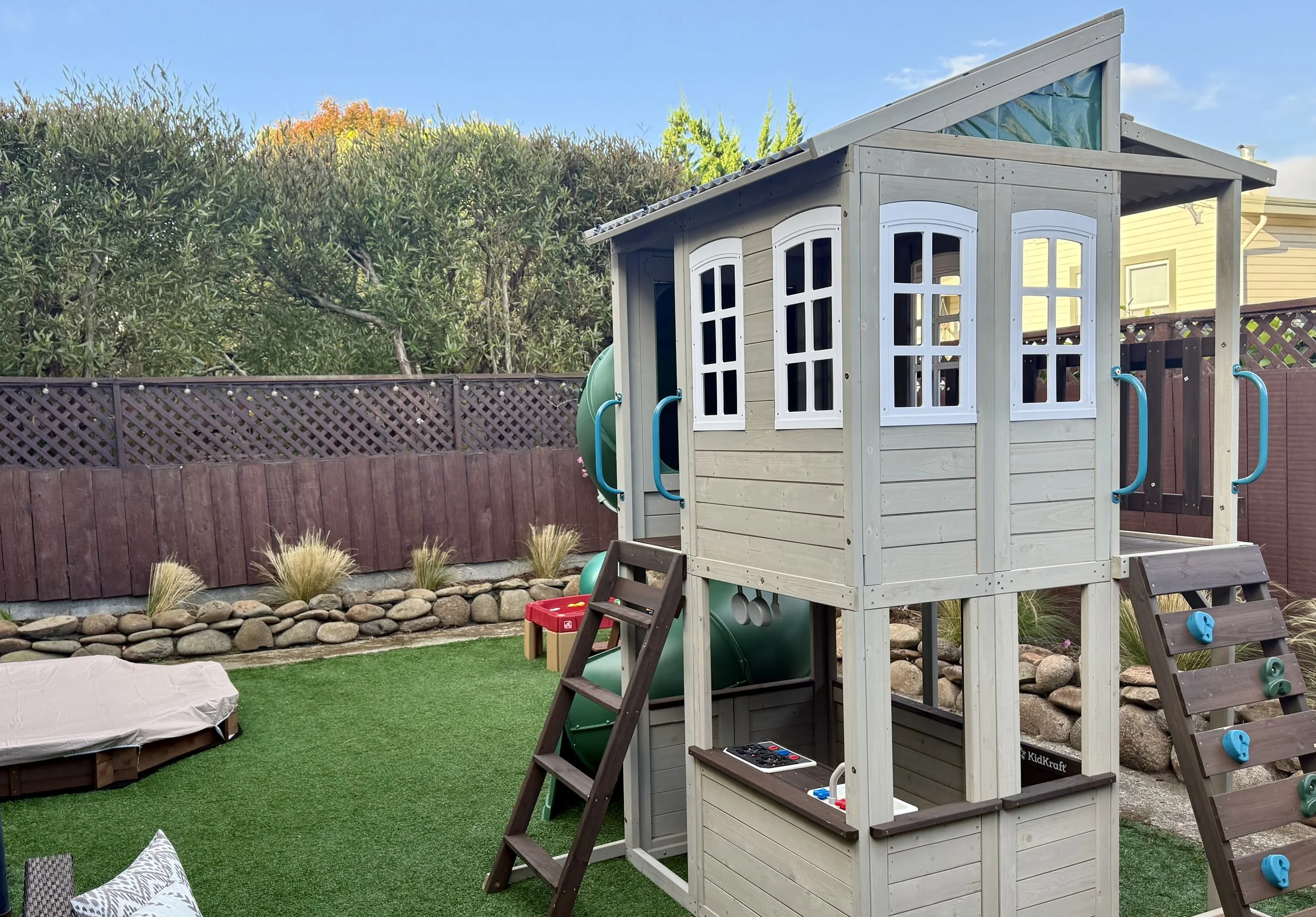 Backyard playground with a wooden playhouse, slide, and climbing wall, surrounded by grass, rocks, and a fence.