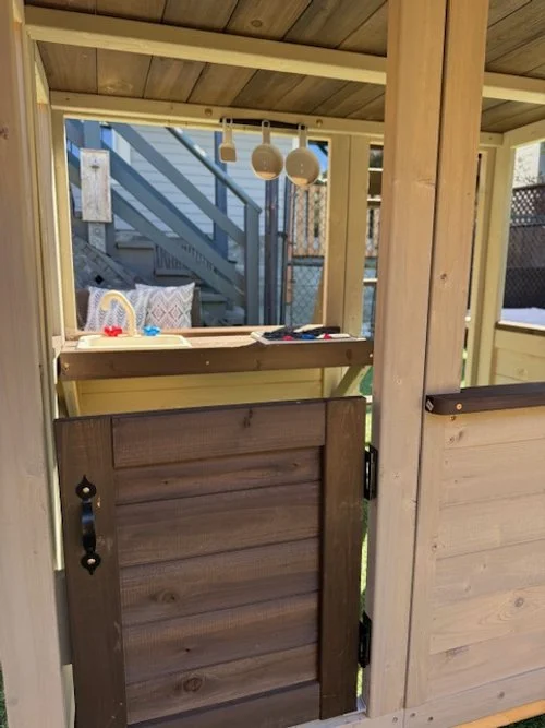 View of a small wooden playhouse with a partial door in the foreground, inside it has a shelf with toys, and two hanging light bulbs. In the background, there is an outdoor wooden staircase and a fence.