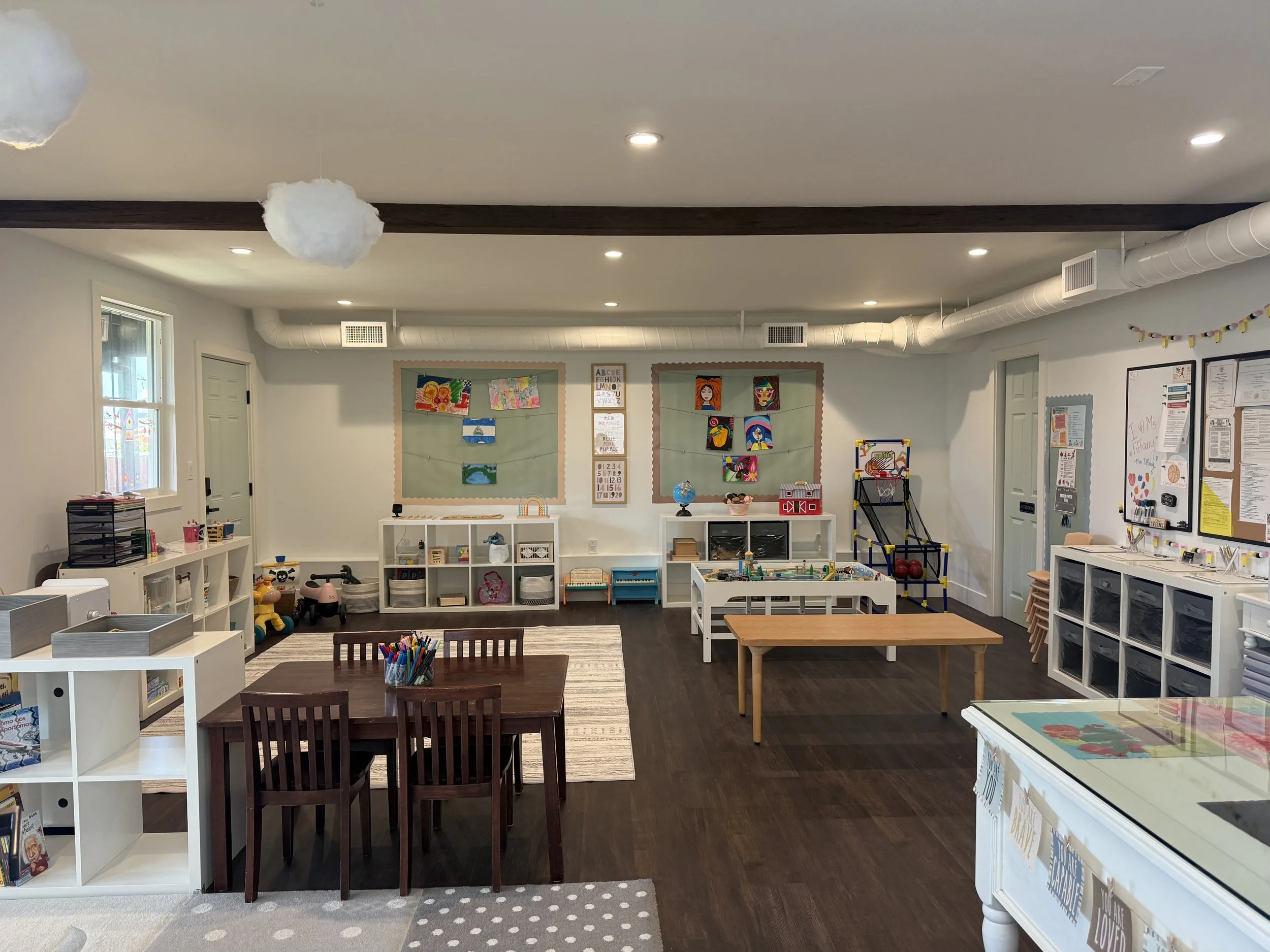 A bright, organized classroom with children's artwork on bulletin boards, small tables and chairs, and shelves with educational toys and supplies.