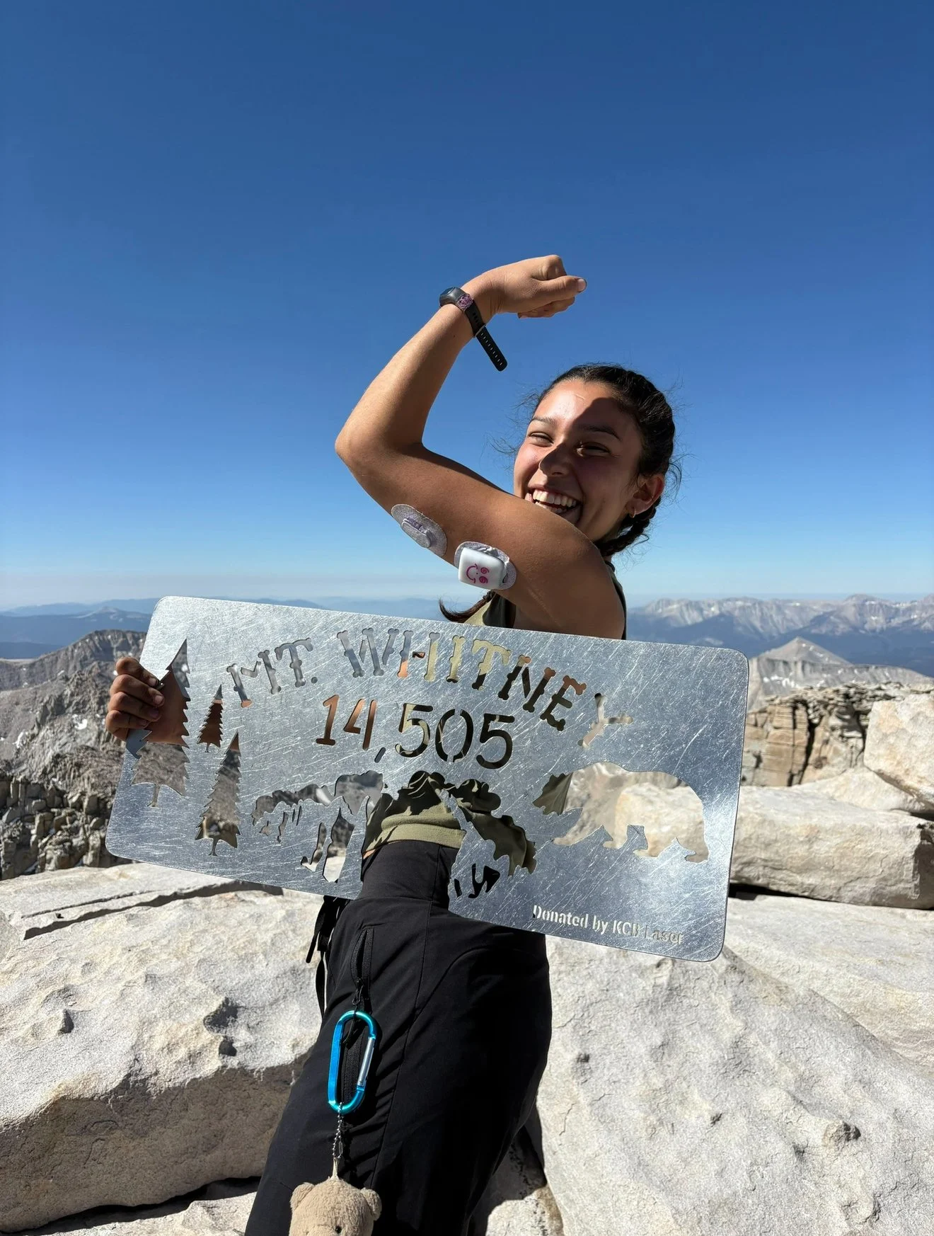 A woman on a mountain summit holding a sign that reads 'Mt. Whitney 14,505', with a mountainous landscape in the background. She is smiling and flexing her arm, wearing a watch and a bandage on her forearm.
