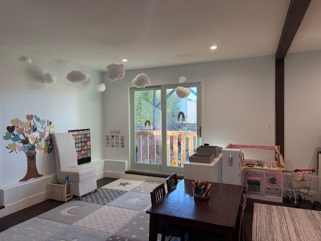 Child's playroom with cloud decorations hanging from ceiling, a sliding glass door leading outside, a colorful tree wall decal, a white armchair, a small play kitchen, a wooden table with chairs, and various toys and storage bins.