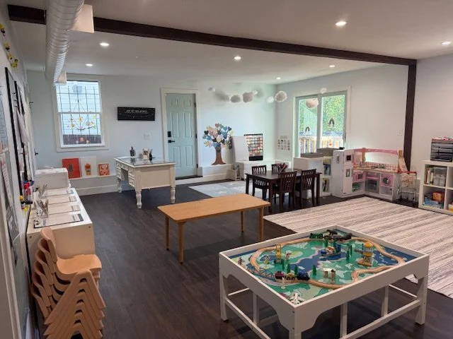 A spacious children's playroom with a train table, a picnic table, a dollhouse, and wall decorations, including a tree mural and hanging clouds, with furniture and toys arranged for play and activities.