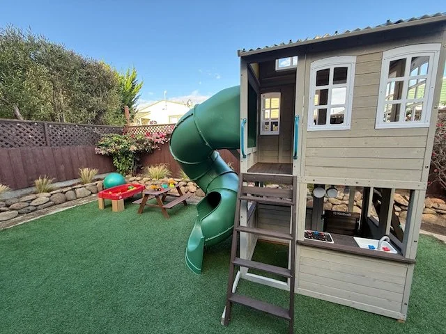 Children's backyard playhouse with a green slide, a small sandbox, and various toys on artificial grass.