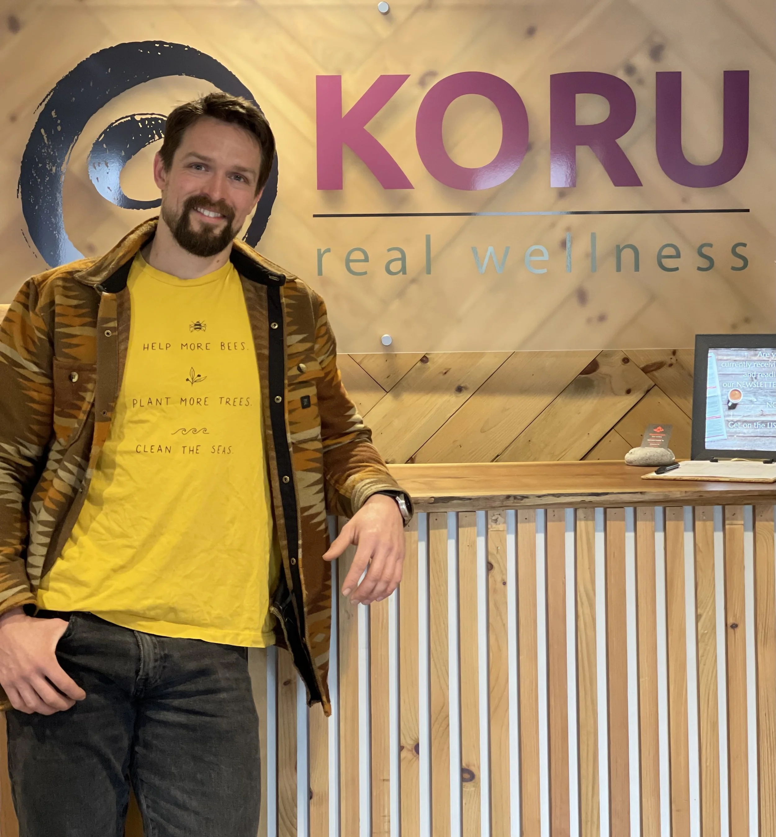 Colin is wearing a yellow shirt with a brown patterned jacket at the front desk in KORU.