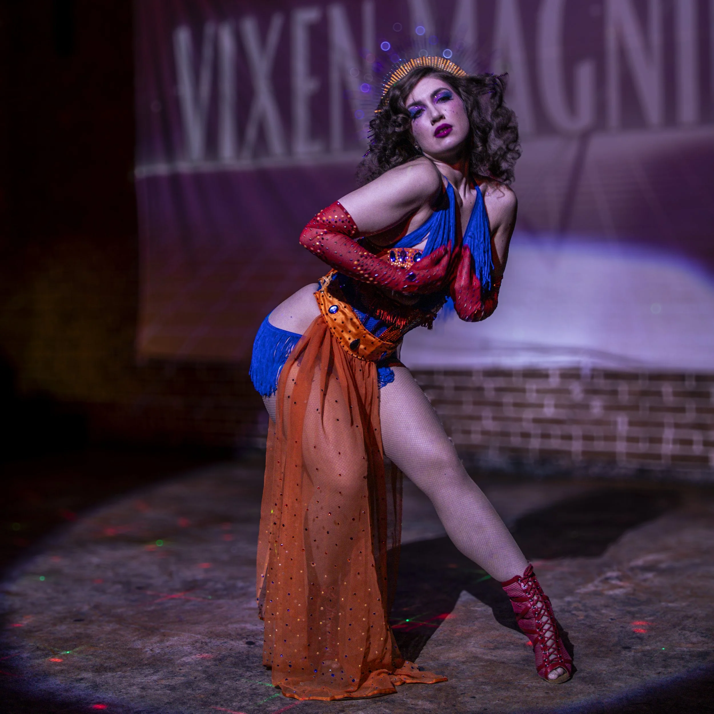 A woman with curly hair performing on stage, wearing a colorful costume with blue, orange, and red elements, including a sheer orange skirt with rhinestones, fishnet tights, and red lace-up heels, with a backdrop displaying the word 'MIXEN'.