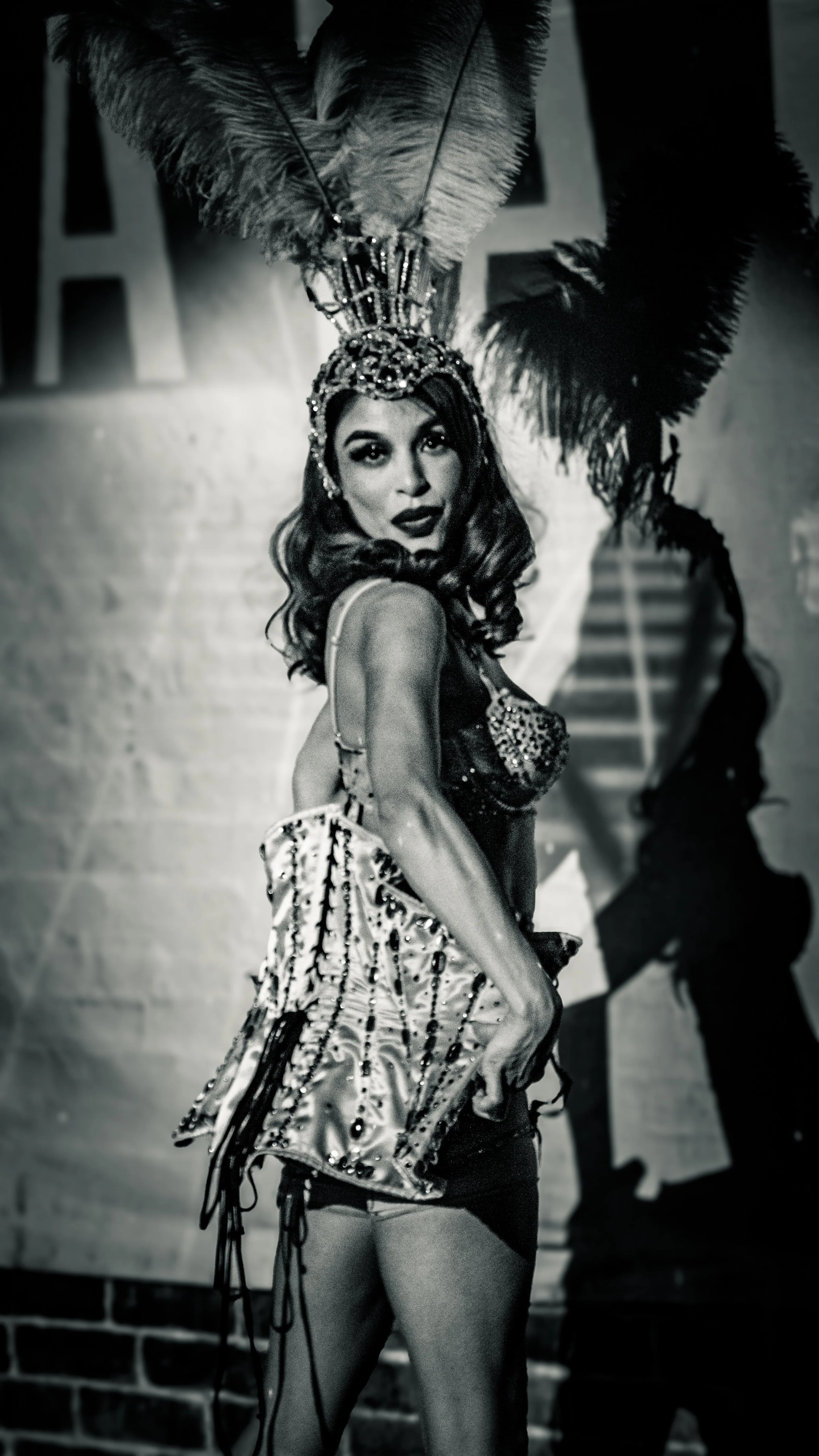 A woman dressed in a showgirl costume with a feathered headdress, sequined top, and short skirt, striking a pose in front of a backdrop with a staircase and the letters 'A'. She has dark hair and makeup, and appears confident.