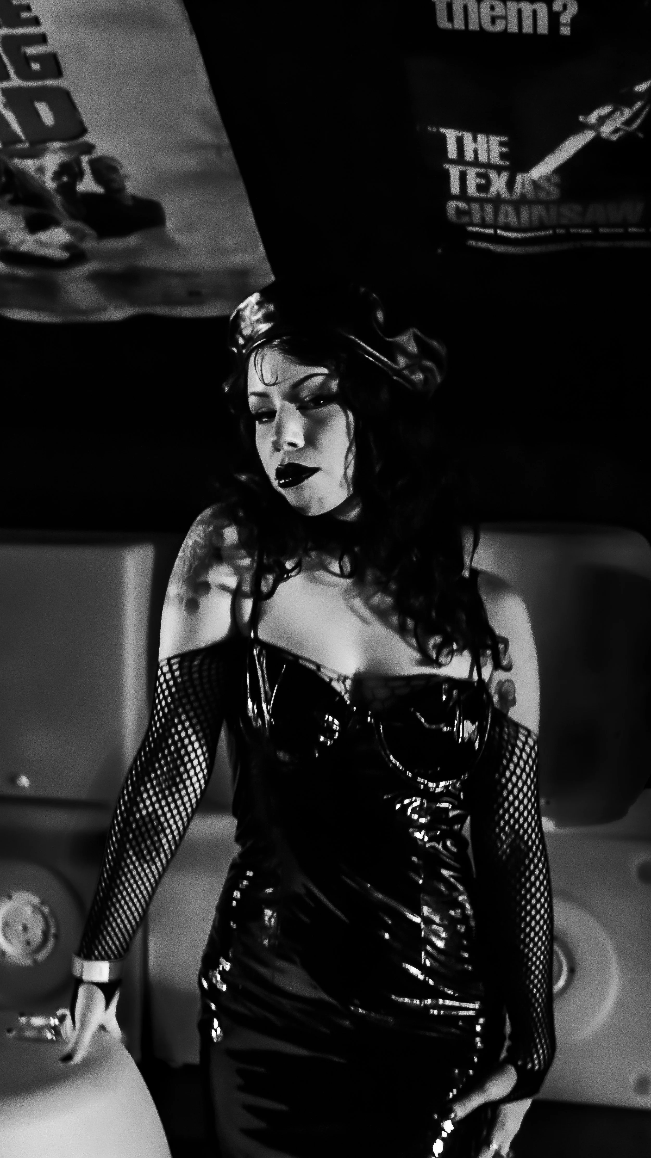 A woman with dark curly hair and dark lipstick wearing a shiny, black, fishnet-sleeved dress, standing indoors.