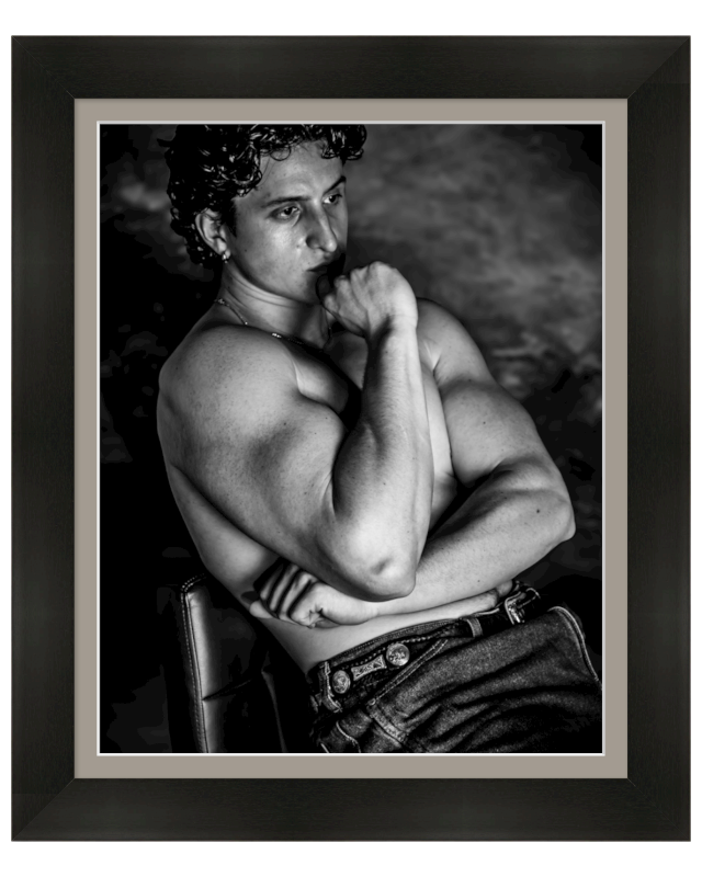 Black and white portrait of a man with short curly hair, sitting topless with arms crossed, resting chin on hand, and looking thoughtfully.