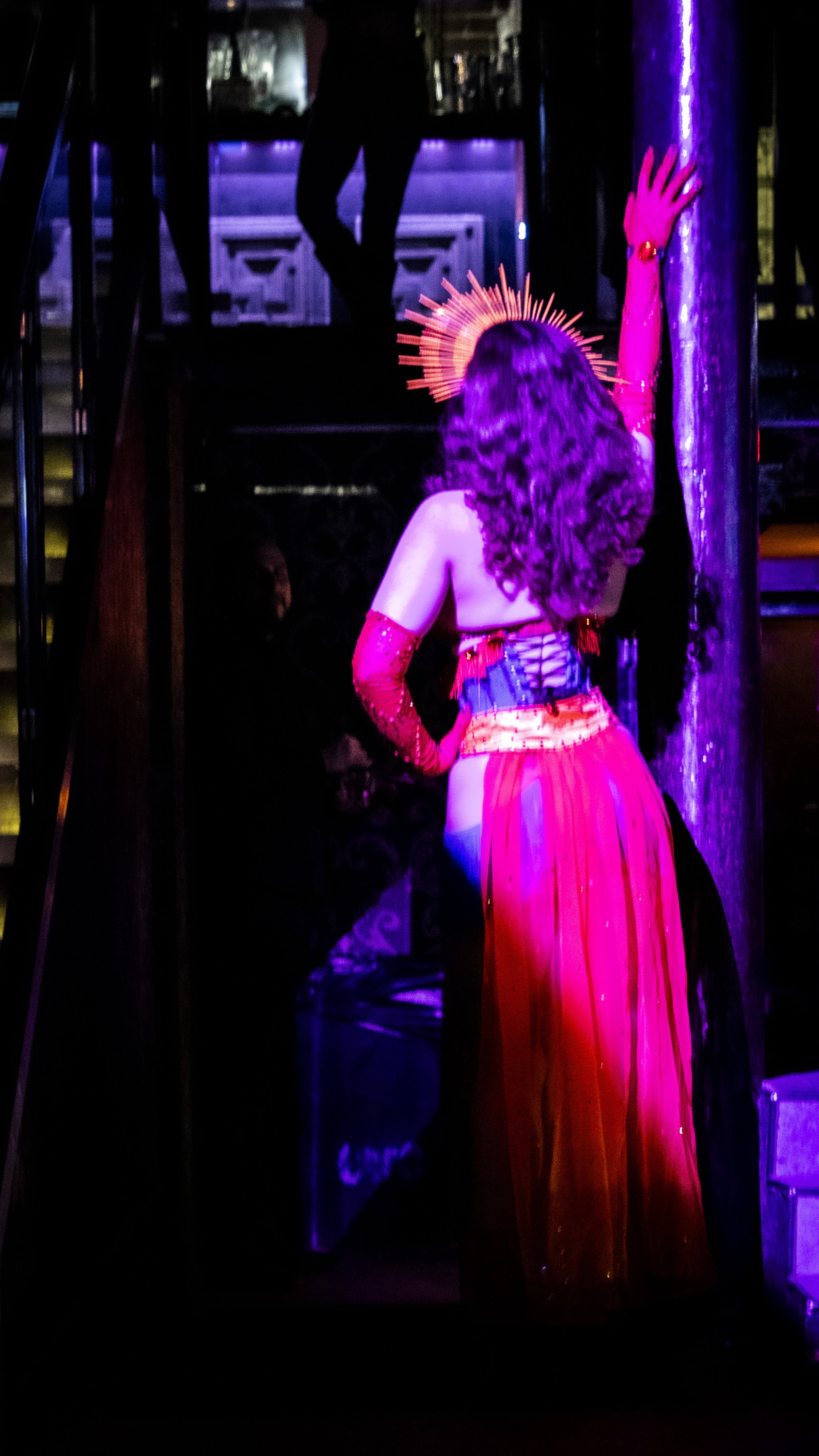 A performer dressed in a red and purple costume with long red gloves, curly hair, and a golden halo-like accessory on her head is posed with her back to the camera, leaning against a metallic pole in a dimly lit environment with vibrant purple and pi