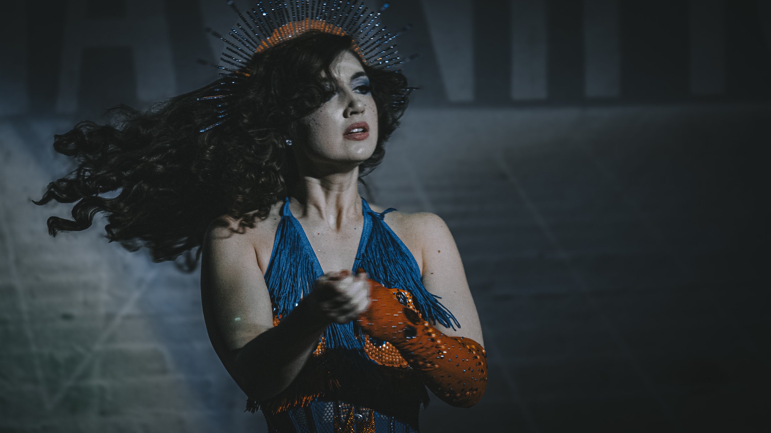A woman with long, curly hair and dramatic makeup, wearing a blue fringed dress and an orange glove with black spots, holding a black-and-orange patterned object, standing in a dark, mysterious environment with a halo-like headpiece.