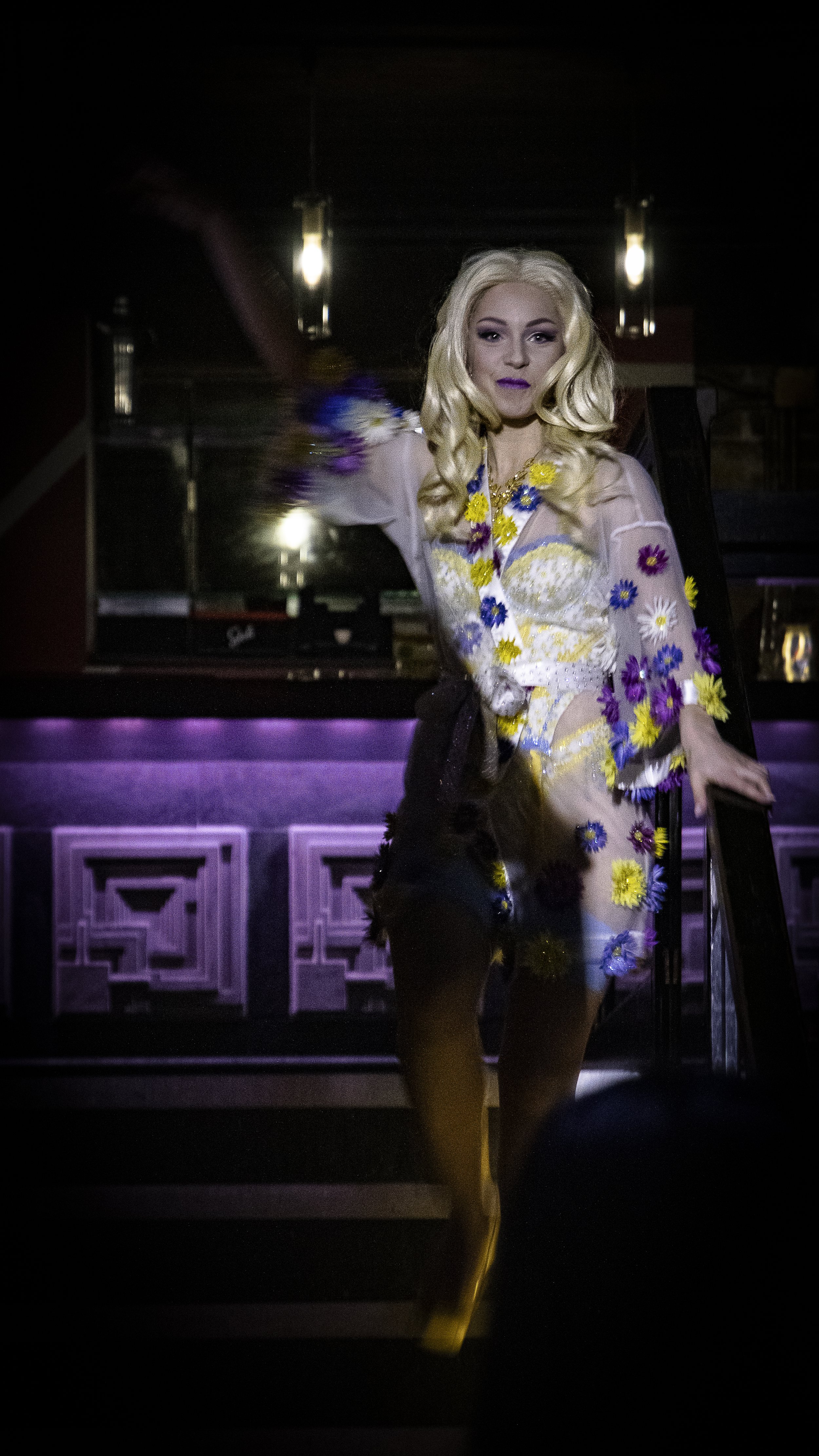 A woman with long blonde hair, wearing a sheer dress decorated with colorful flowers, standing on a staircase in a dimly lit room with purple lighting.