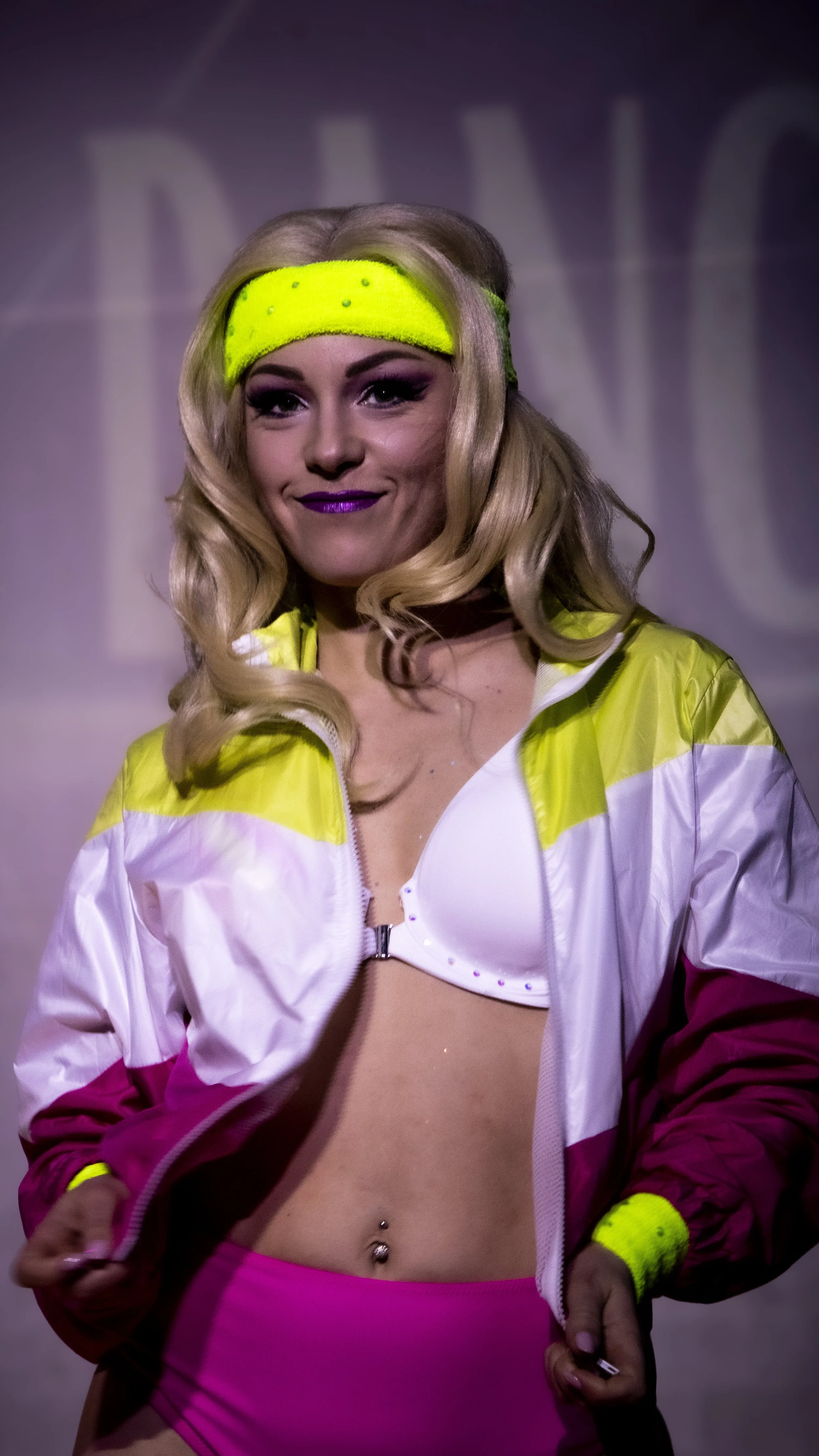 Woman wearing a neon yellow headband, white and pink jacket, white bra, and pink shorts, smiling at the camera.