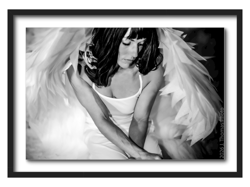 A woman with dark hair and bangs wearing a white dress, sitting with her eyes closed, surrounded by large feathered wings.
