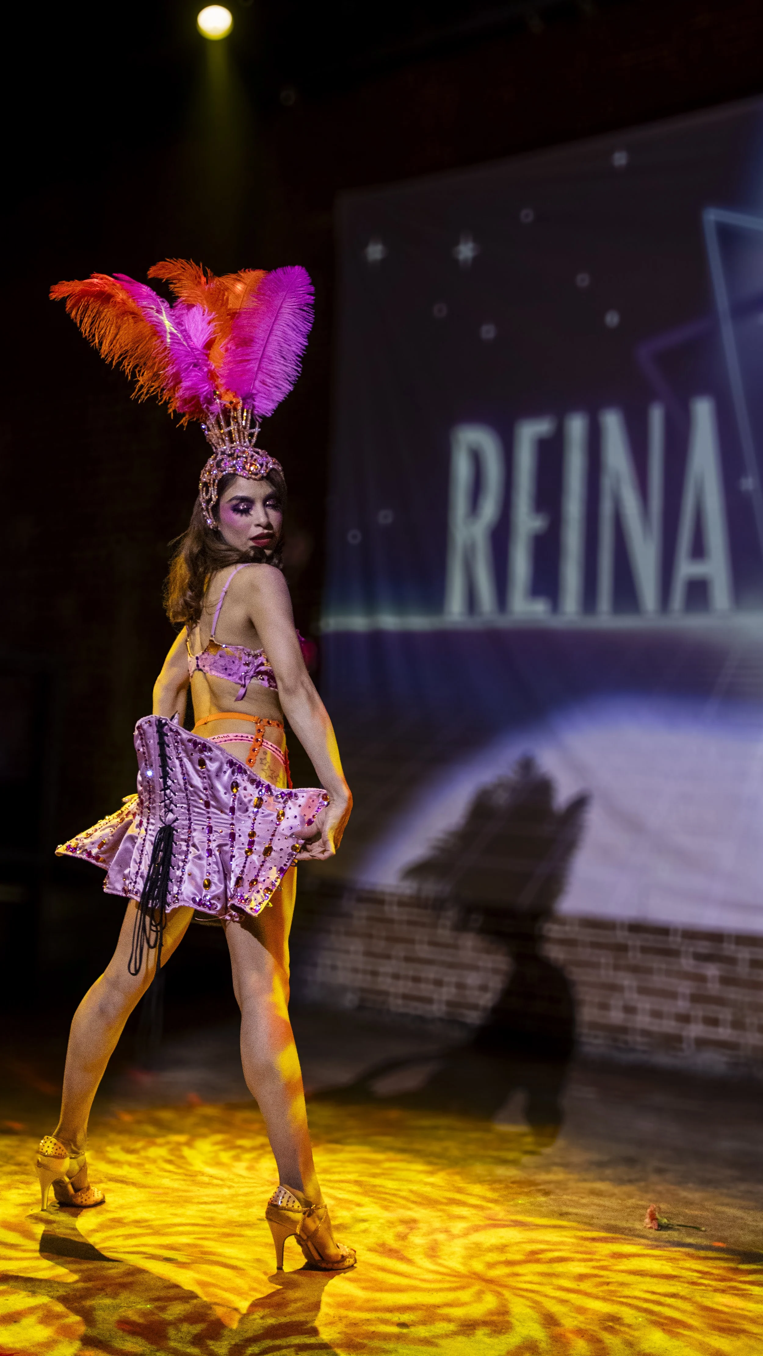 A woman dressed in a pink and purple showgirl costume with a large feathered headdress on stage, holding a matching costume piece, with a backdrop displaying the name 'REINA'.