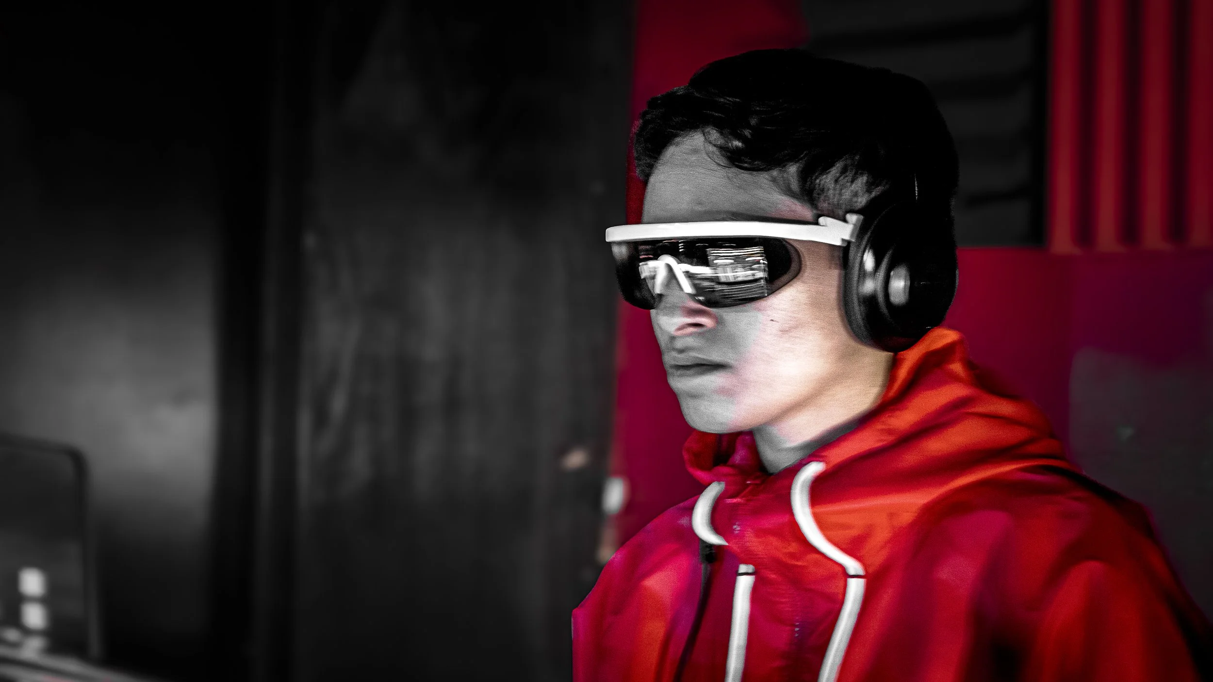A man wearing a red jacket, large black headphones, and sunglasses, working on a computer in a dark room with a red background.