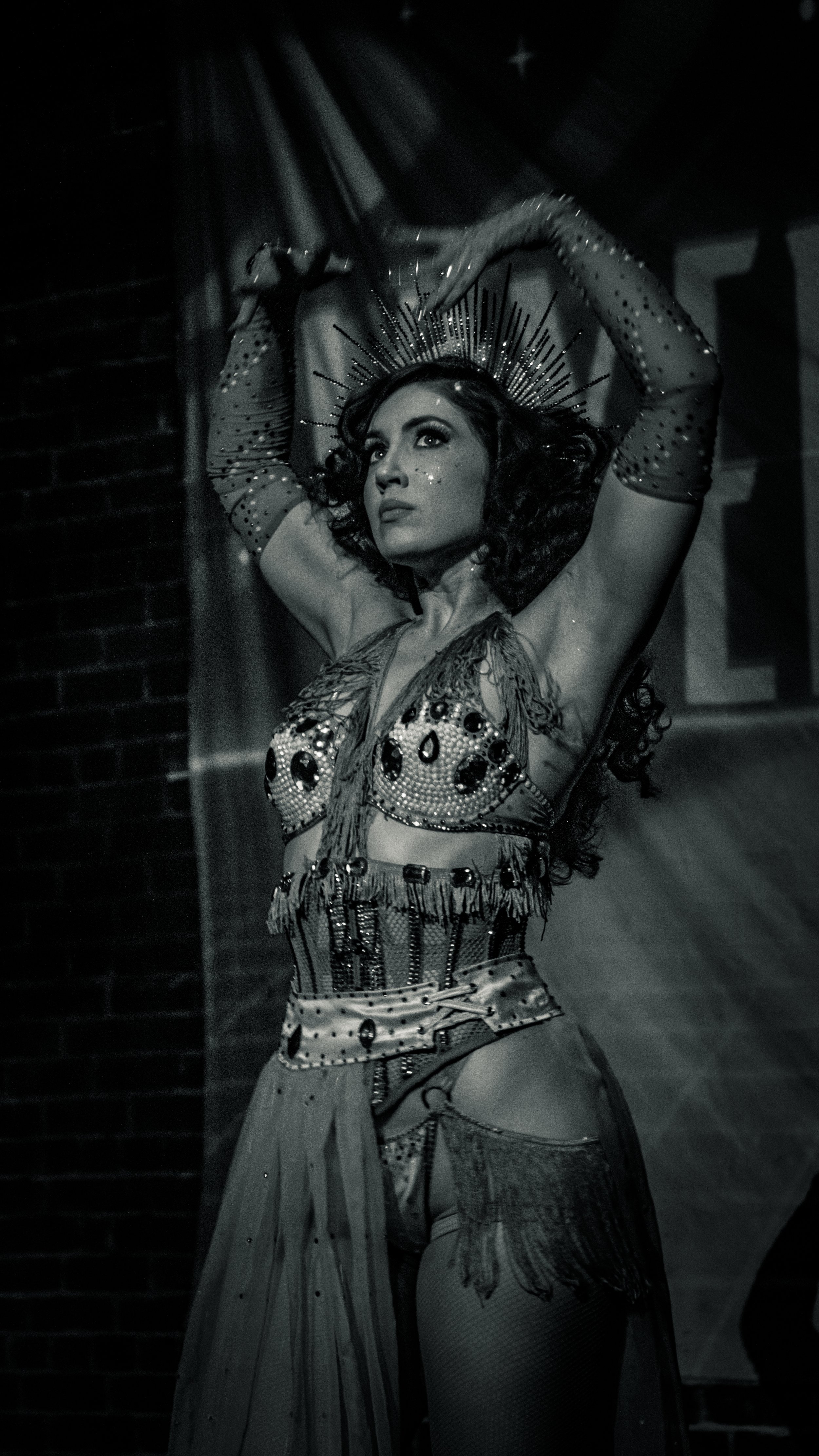 A woman in an elaborate costume with a headdress, glitter makeup, and a beaded top, performing or posing with arms raised.