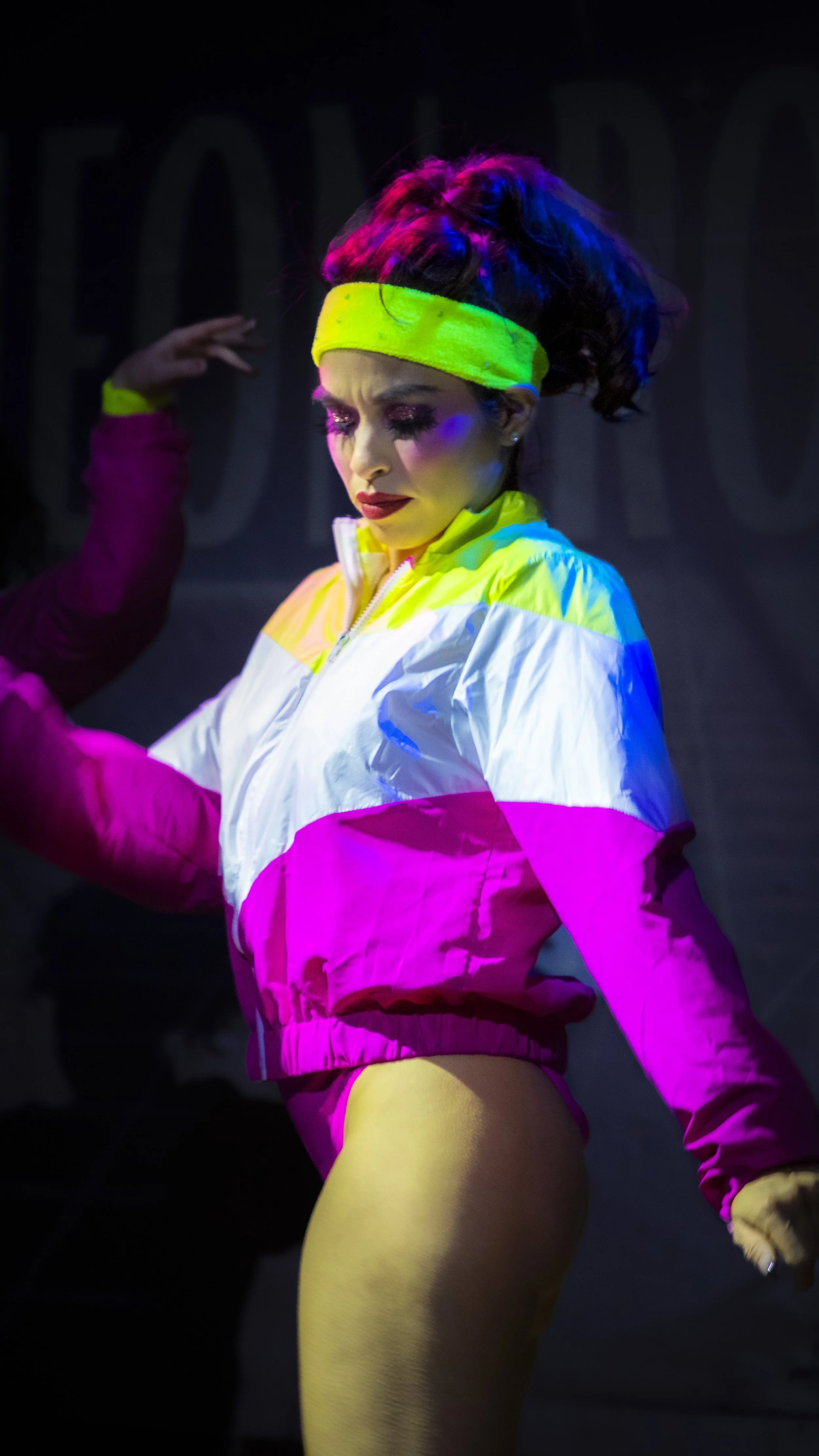 A woman with colorful makeup and a vibrant, retro-style workout outfit, including a yellow headband, a multicolored jacket, and yellow leggings, is dancing or performing on stage with a dark background and colorful lighting.