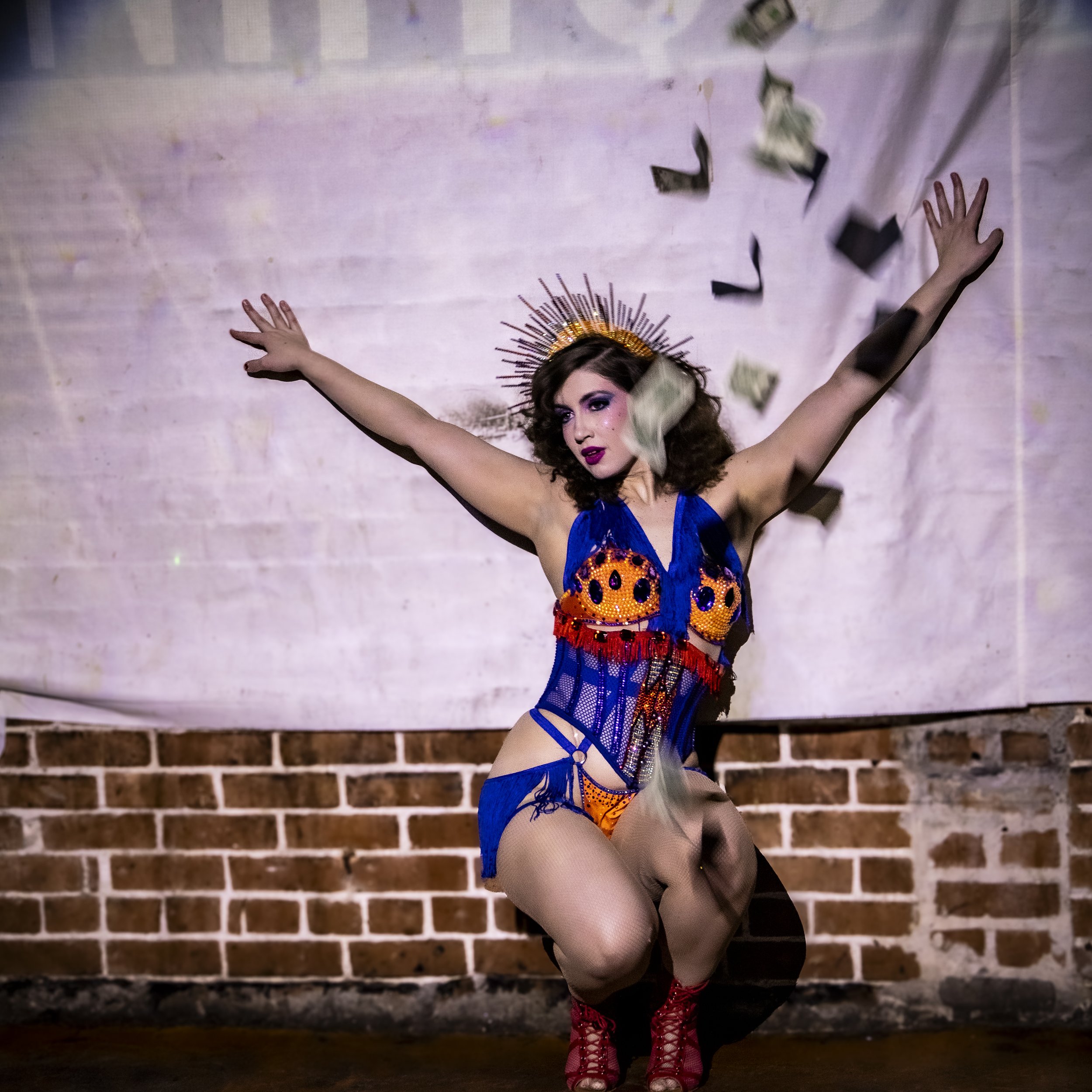 A performer crouching with arms outstretched, wearing a colorful costume with a crown and performing on stage with falling paper currency in the background.