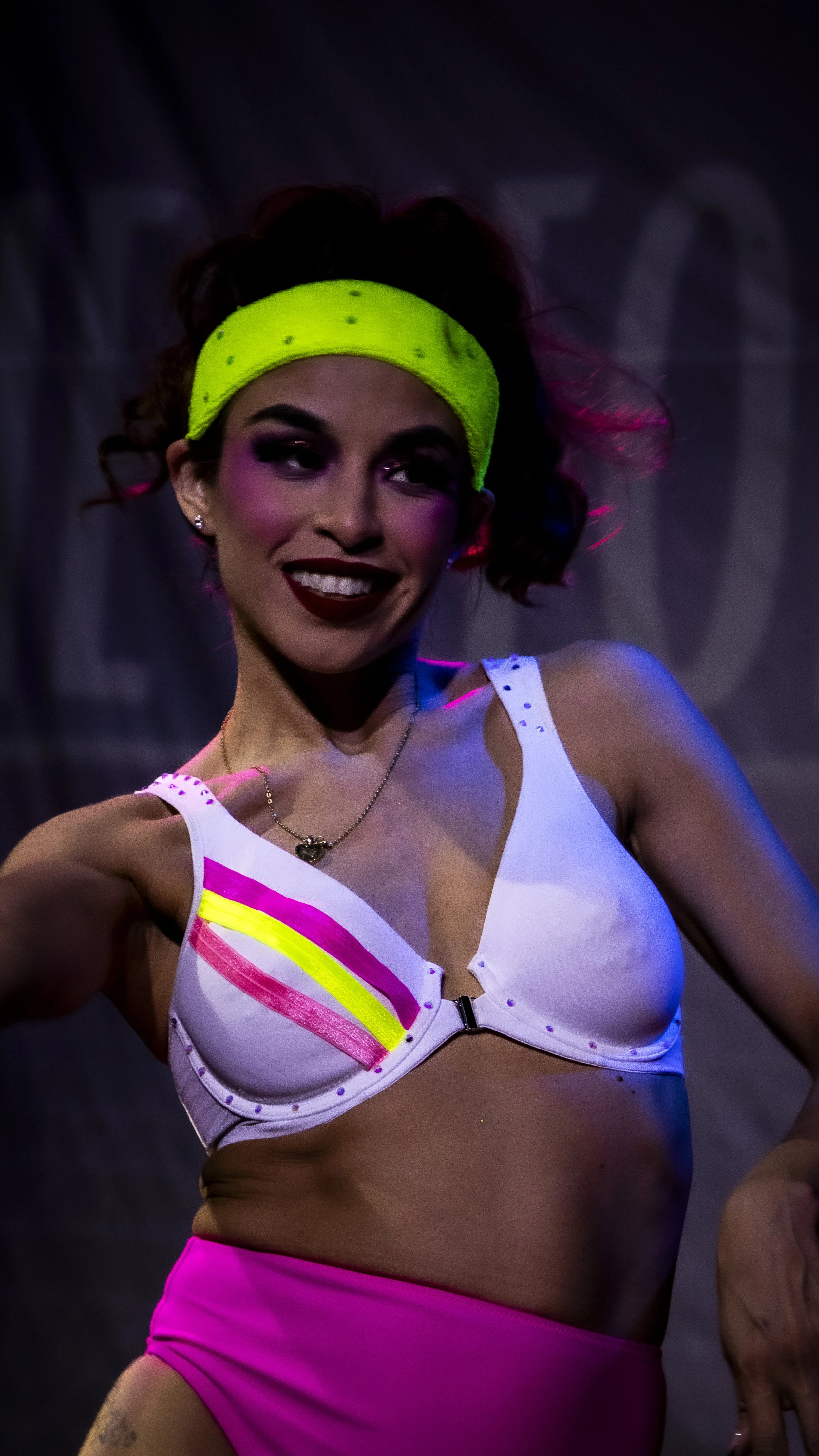 A woman with short curly hair, wearing a neon green headband, a white top with colorful neon stripes, and pink shorts, posing confidently.