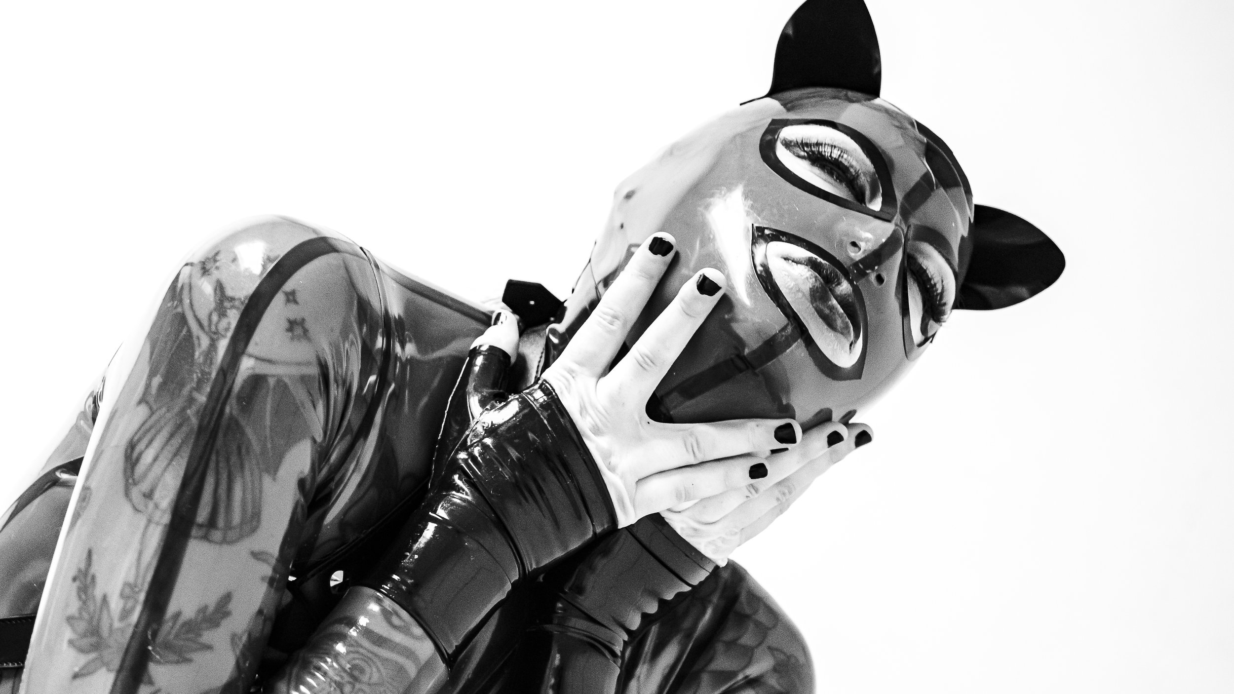 Black and white photo of a person wearing a cat mask and wearing dark latex gloves, covering her face with both hands.