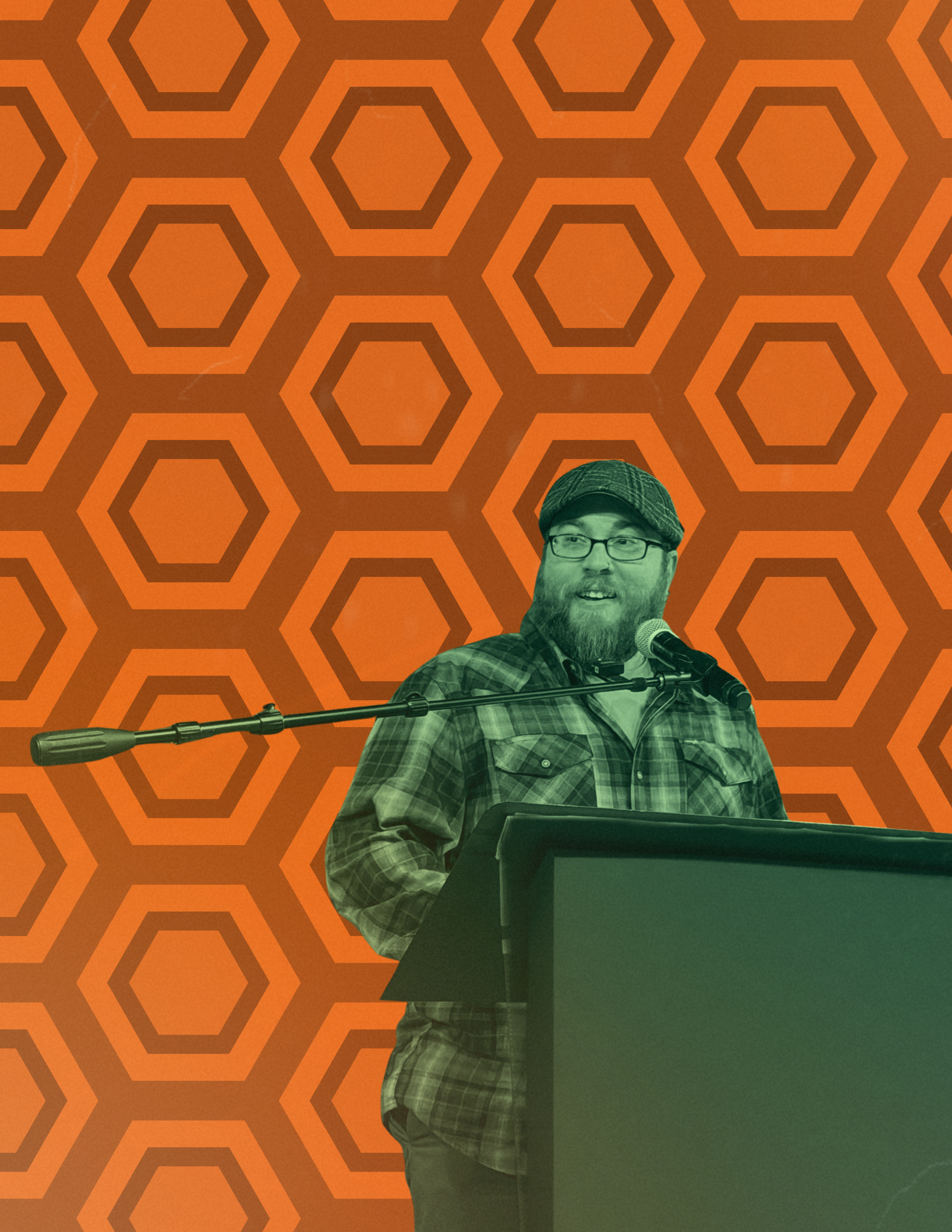 A man with glasses, a beard, and a cap is standing behind a podium, speaking into a microphone against a background of orange honeycomb pattern.
