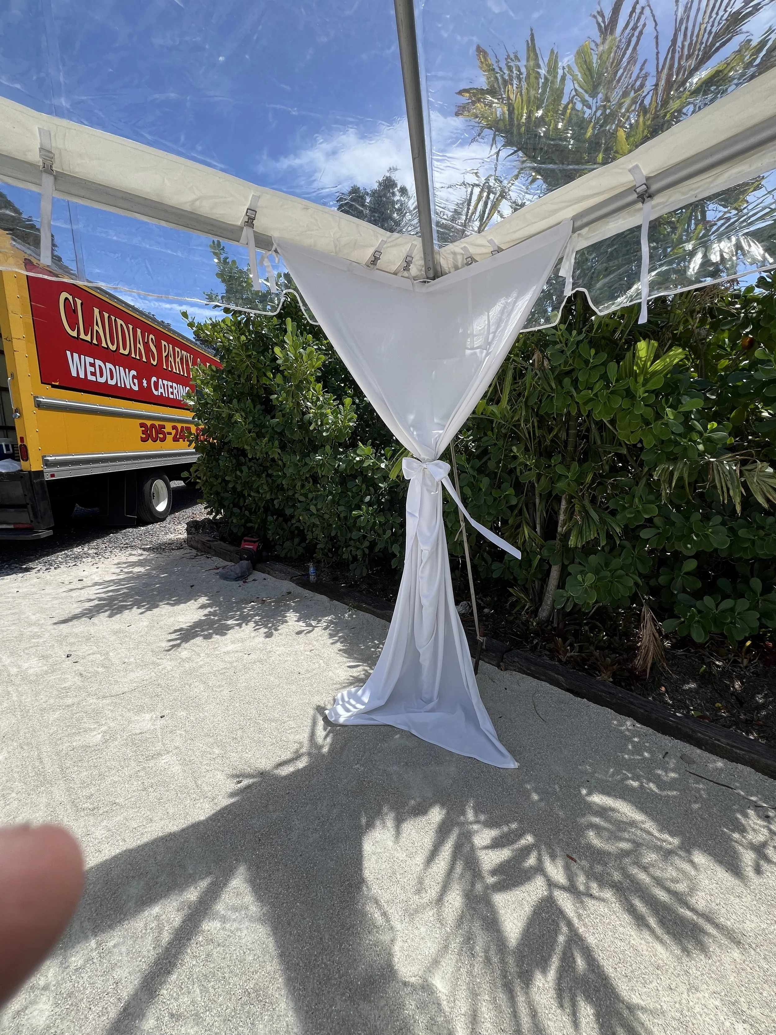 White fabric draped and tied on a pole at an outdoor event, with greenery and a truck in the background under a partly cloudy sky.