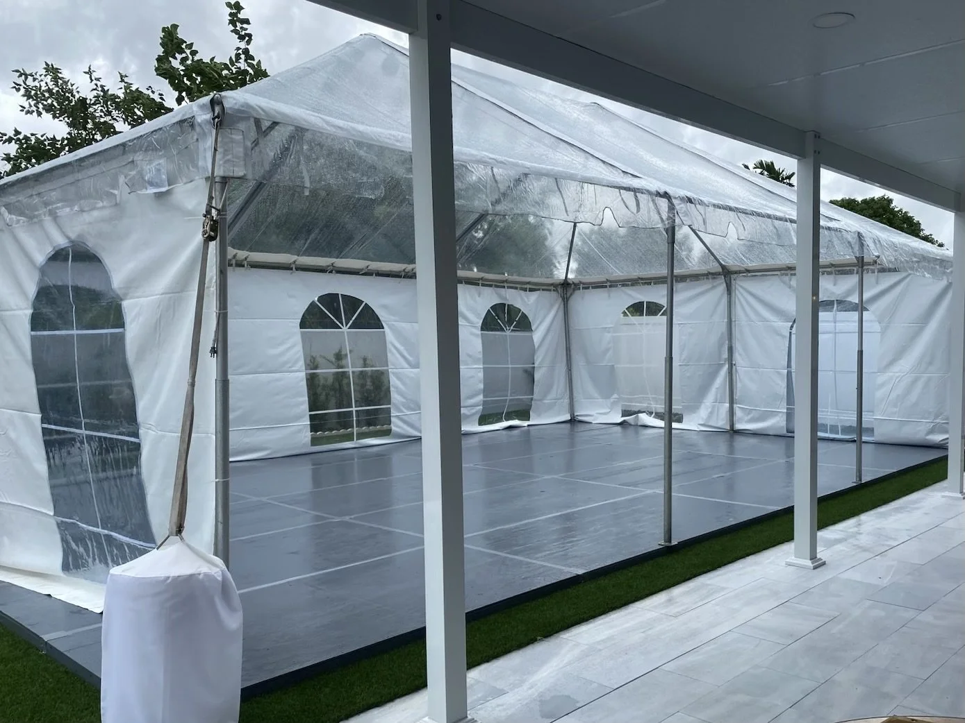 Empty outdoor event tent with transparent plastic walls and arched windows, set up on a patio area with a grassy border, under cloudy skies.