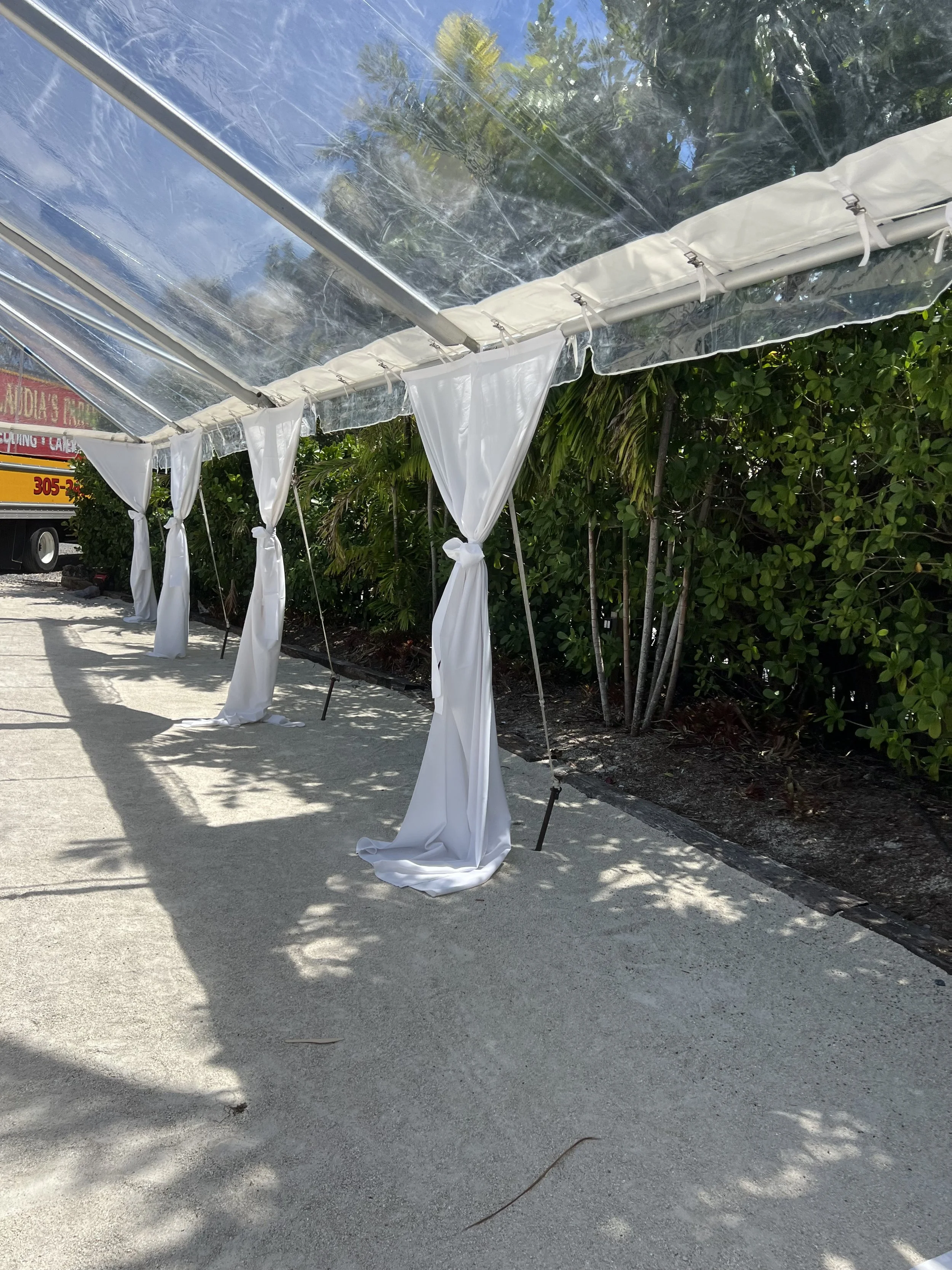 A white fabric canopy with sheer drapes tied back along a concrete sidewalk, surrounded by green bushes and trees, with a truck partially visible in the background.